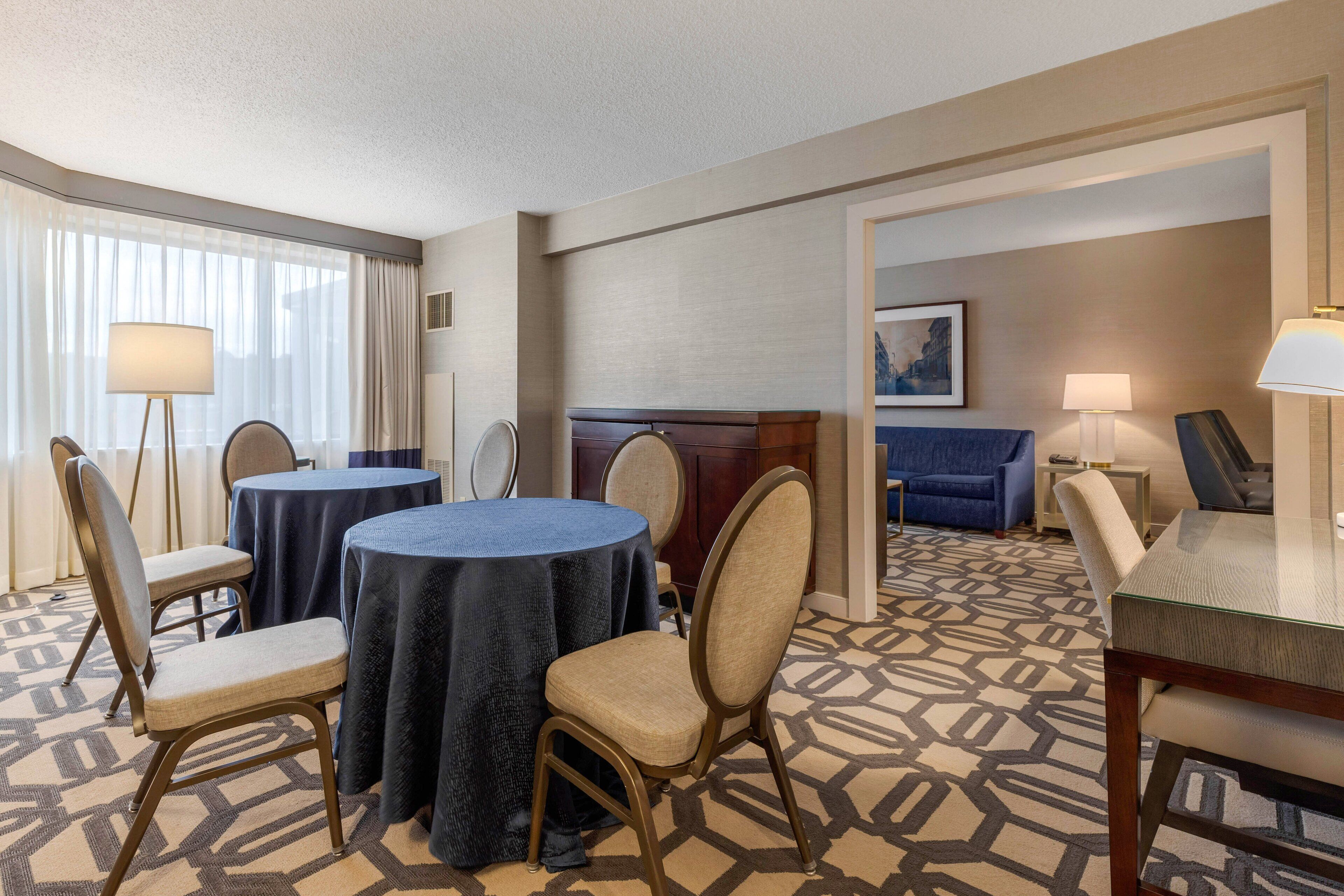 A hotel room with a dining area featuring round tables and chairs, a bed, a sofa, and a lamp.
