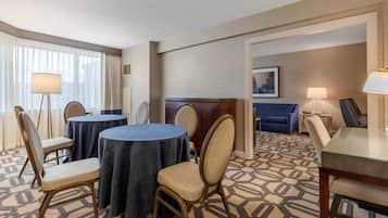 A hotel room with a dining area featuring round tables and chairs, a bed, a sofa, and a lamp.