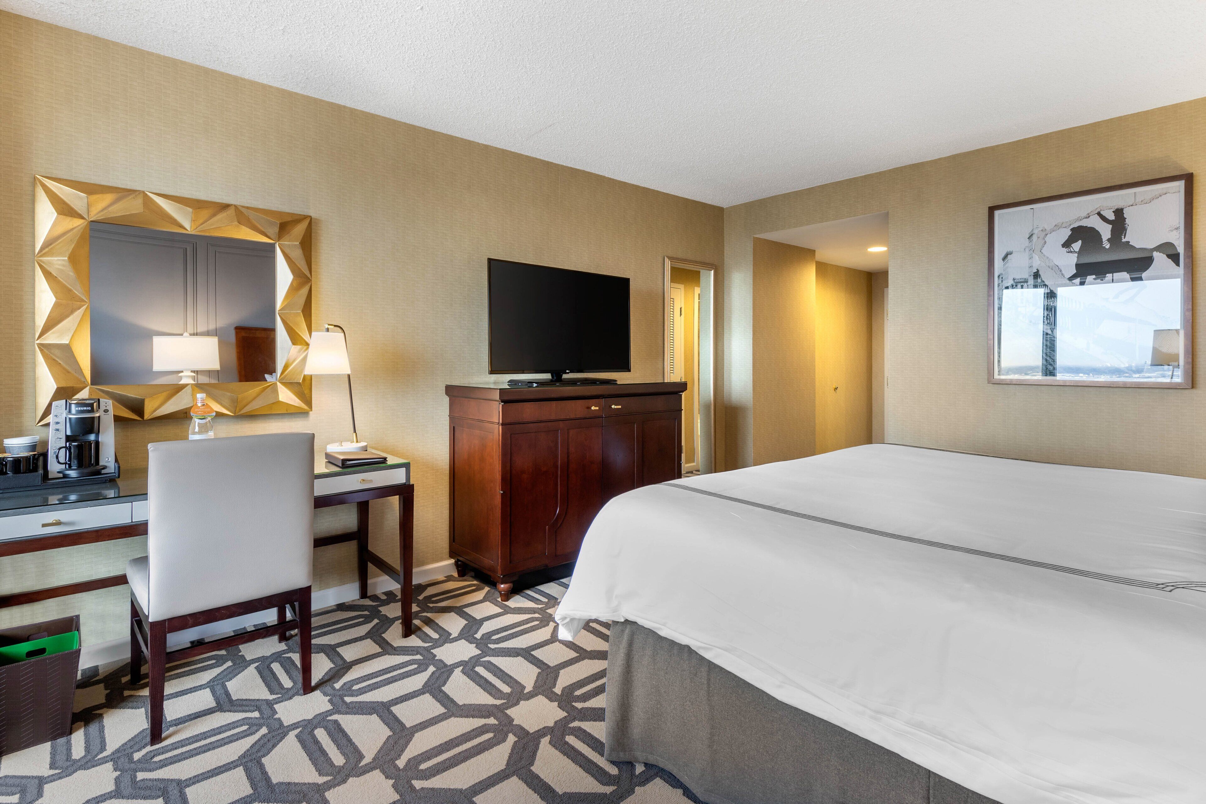 A hotel room with a bed, desk, chair, TV, and a large mirror.