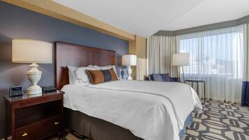 A hotel room with a large bed, two bedside tables with lamps, a nightstand, and a view of the city through large windows.
