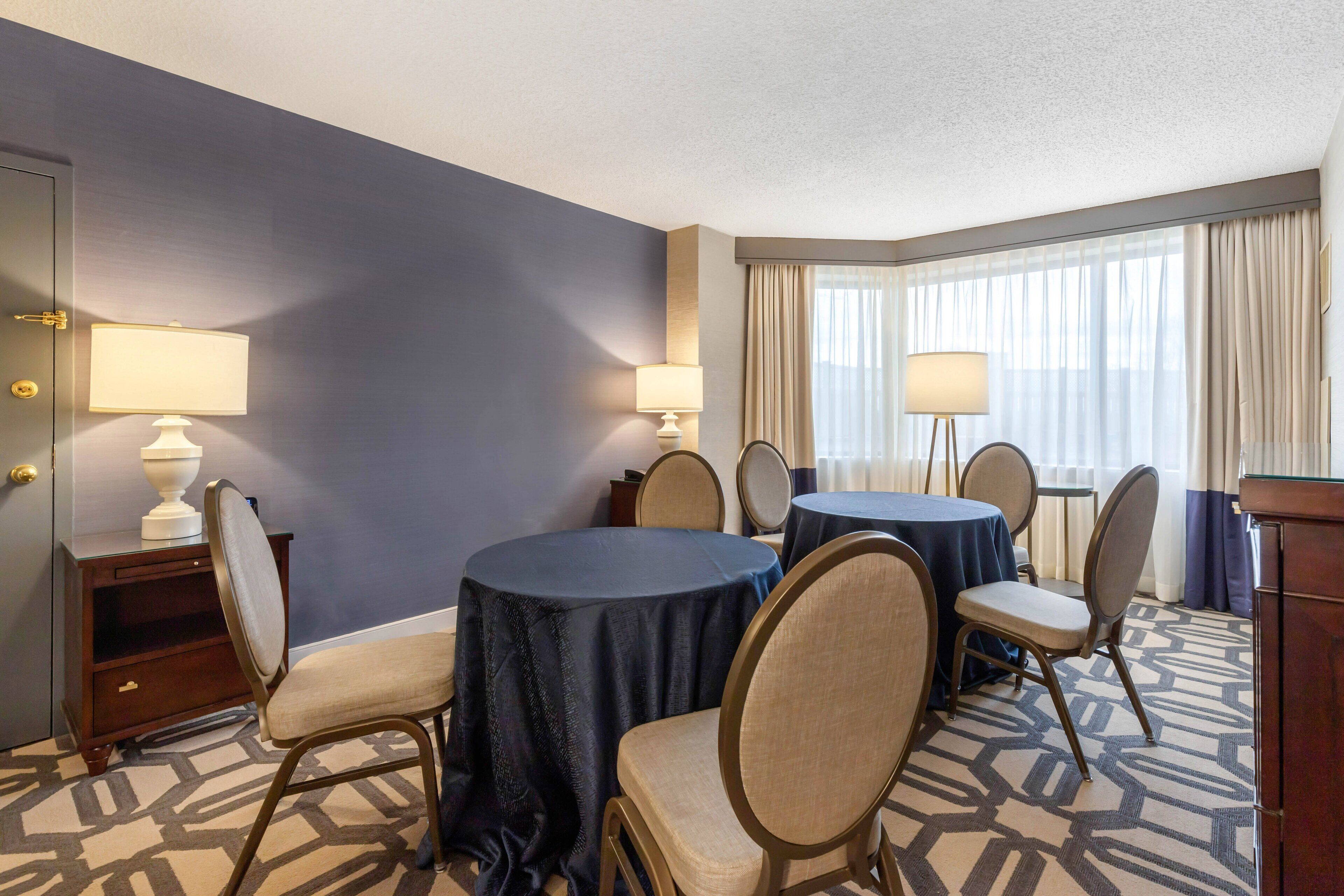 A hotel room with a dining area featuring round tables and chairs, a small side table with a lamp, and a large window with curtains.