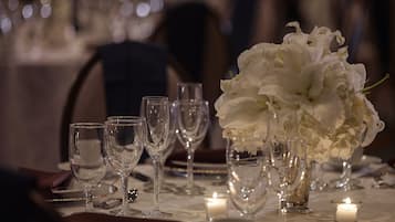 Una mesa lista para cenar, con copas de vino, una vela y un florero con flores blancas.