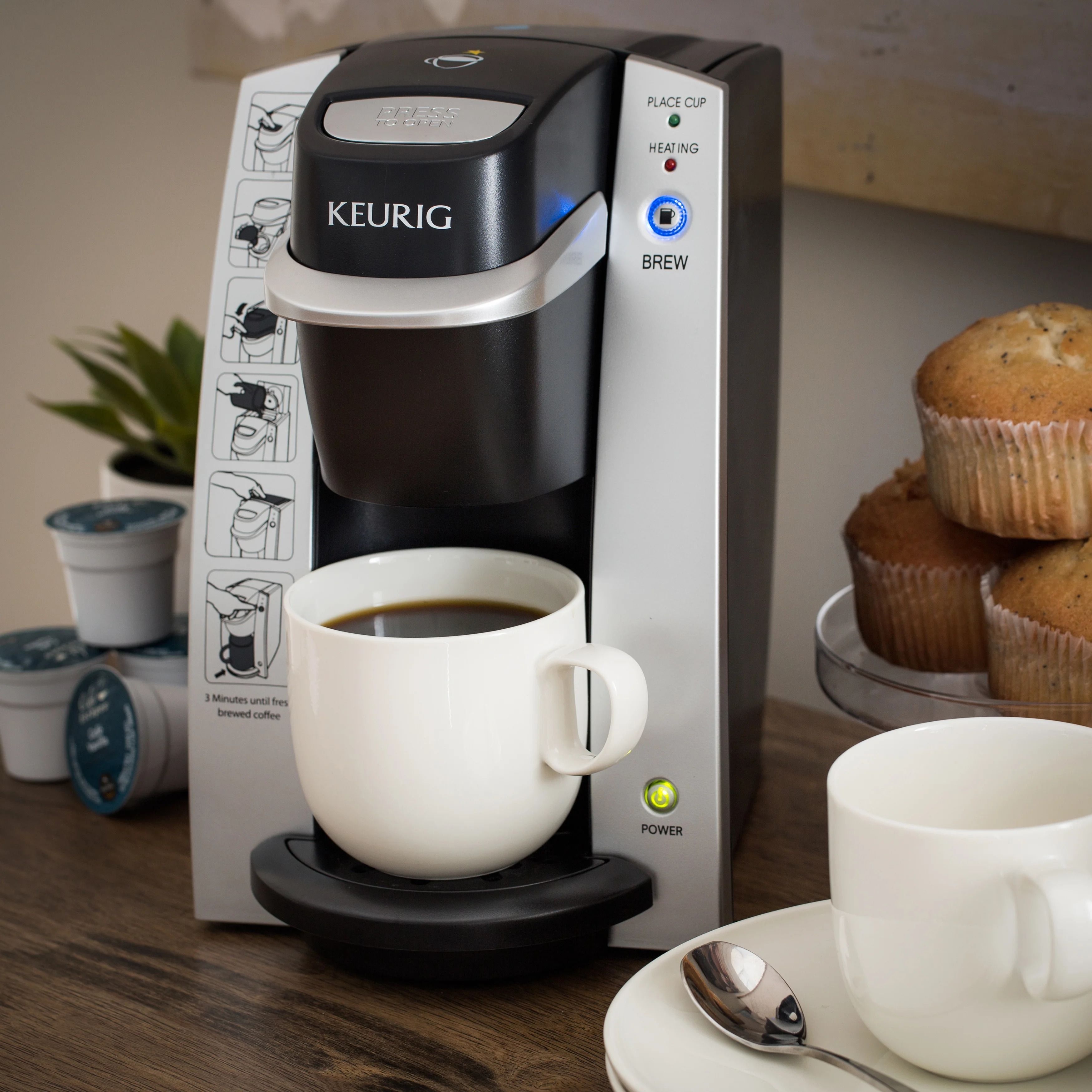 A Keurig coffee maker with a cup of coffee on its drip tray, a muffin in the background, and a spoon on a saucer.