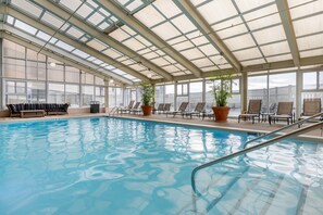 Una piscina interior con techos de vidrio transparente, sillones de descanso y plantas en macetas.