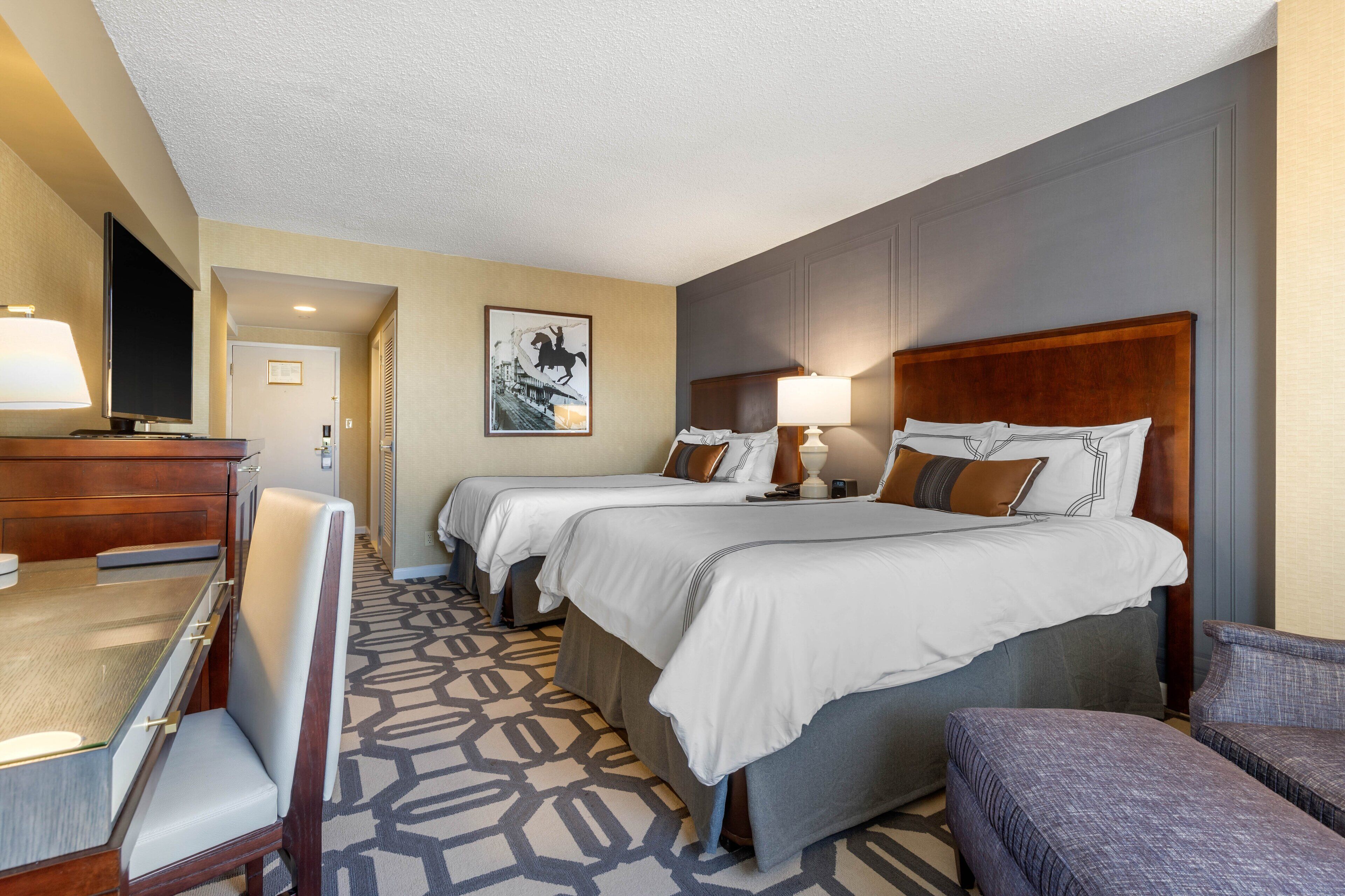 A hotel room with two beds, a desk, a chair, and a television.