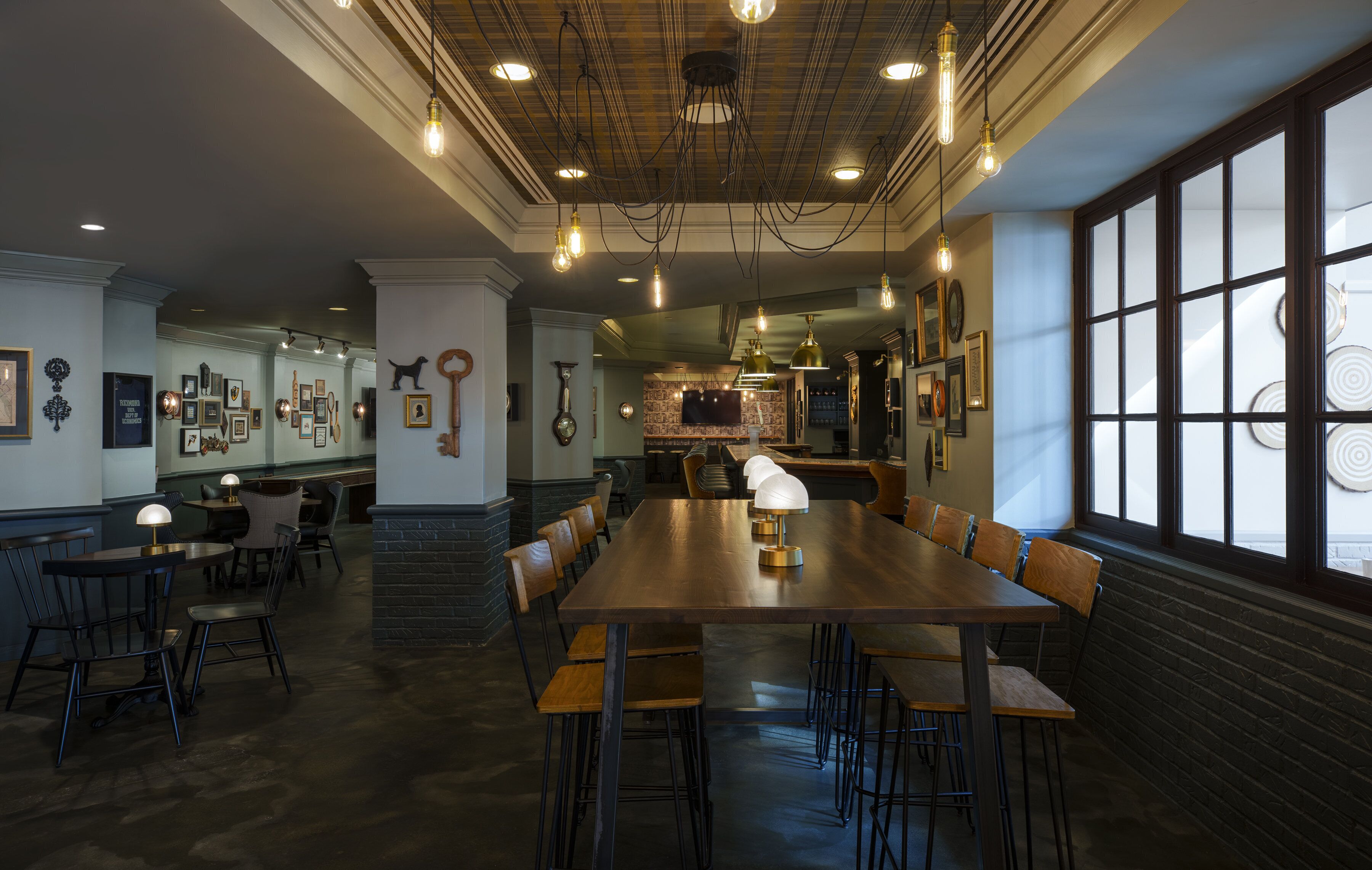Un vaste espace dédié à la salle à manger, avec une longue table en bois, de nombreuses chaises et des luminaires suspendus.