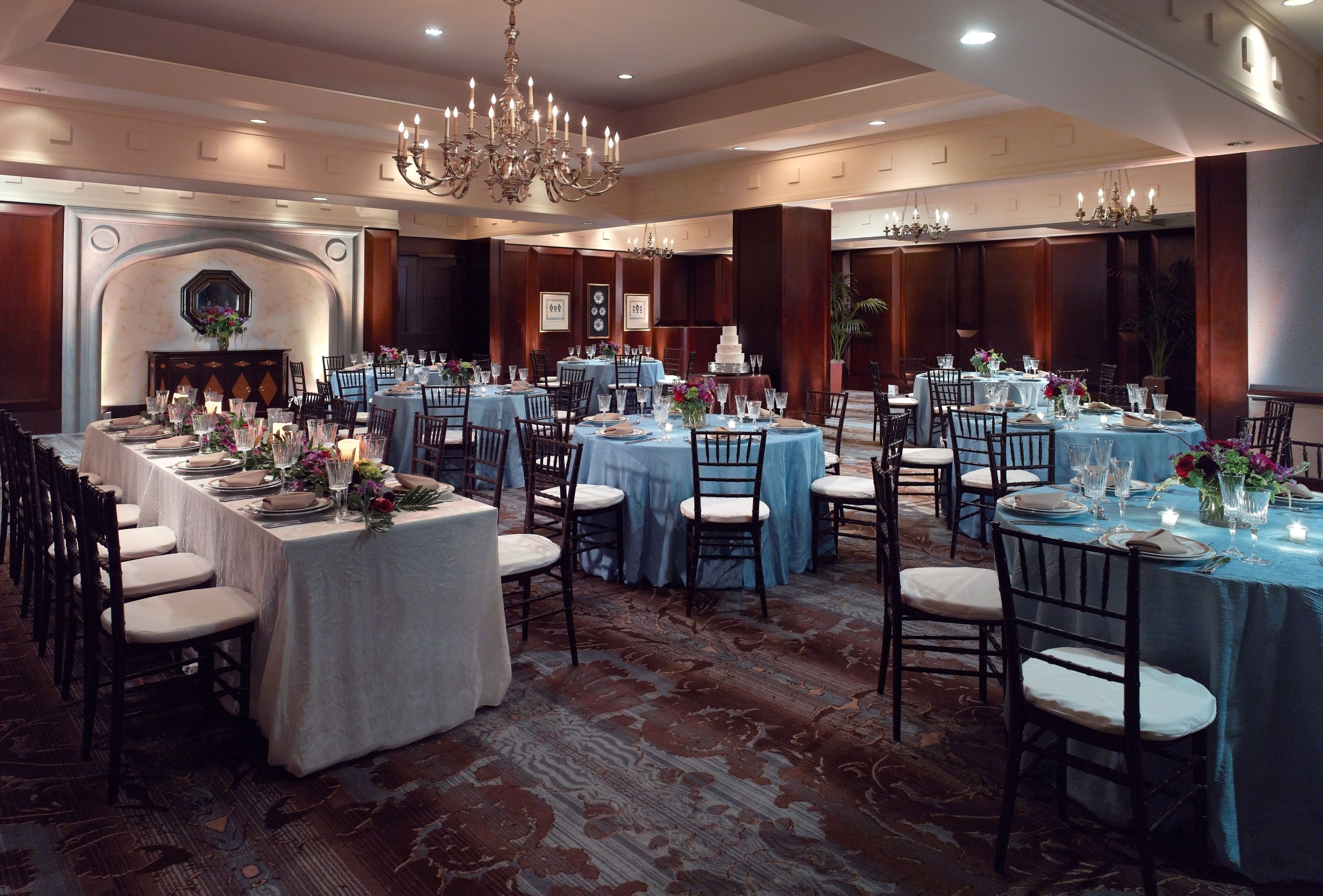 A banquet hall with multiple round tables, each set with chairs and tablecloths, and a large chandelier hanging from the ceiling.