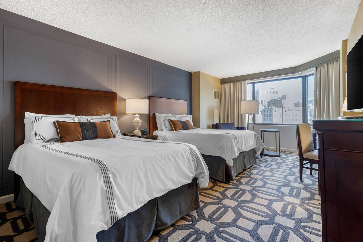 Een hotelkamer met twee bedden, een bureau, een stoel en een groot raam met uitzicht op de stad.