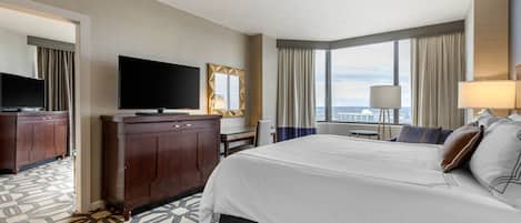 A hotel room with a large bed, a flat-screen TV, a desk, and a view of the city through large windows.