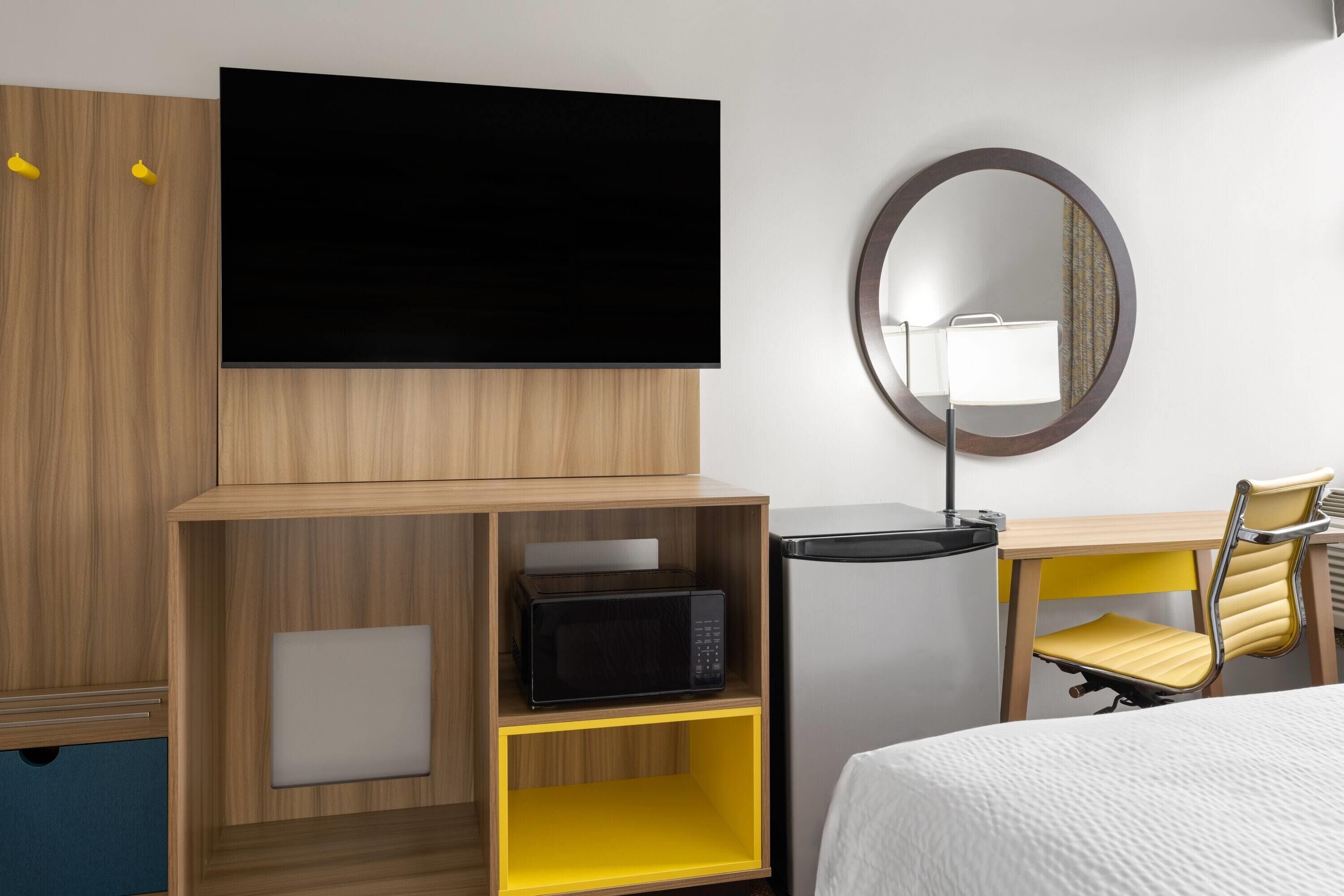 A hotel room with a flat-screen TV mounted on the wall, a microwave on a wooden shelf, a desk with a chair, and a round mirror on the wall.