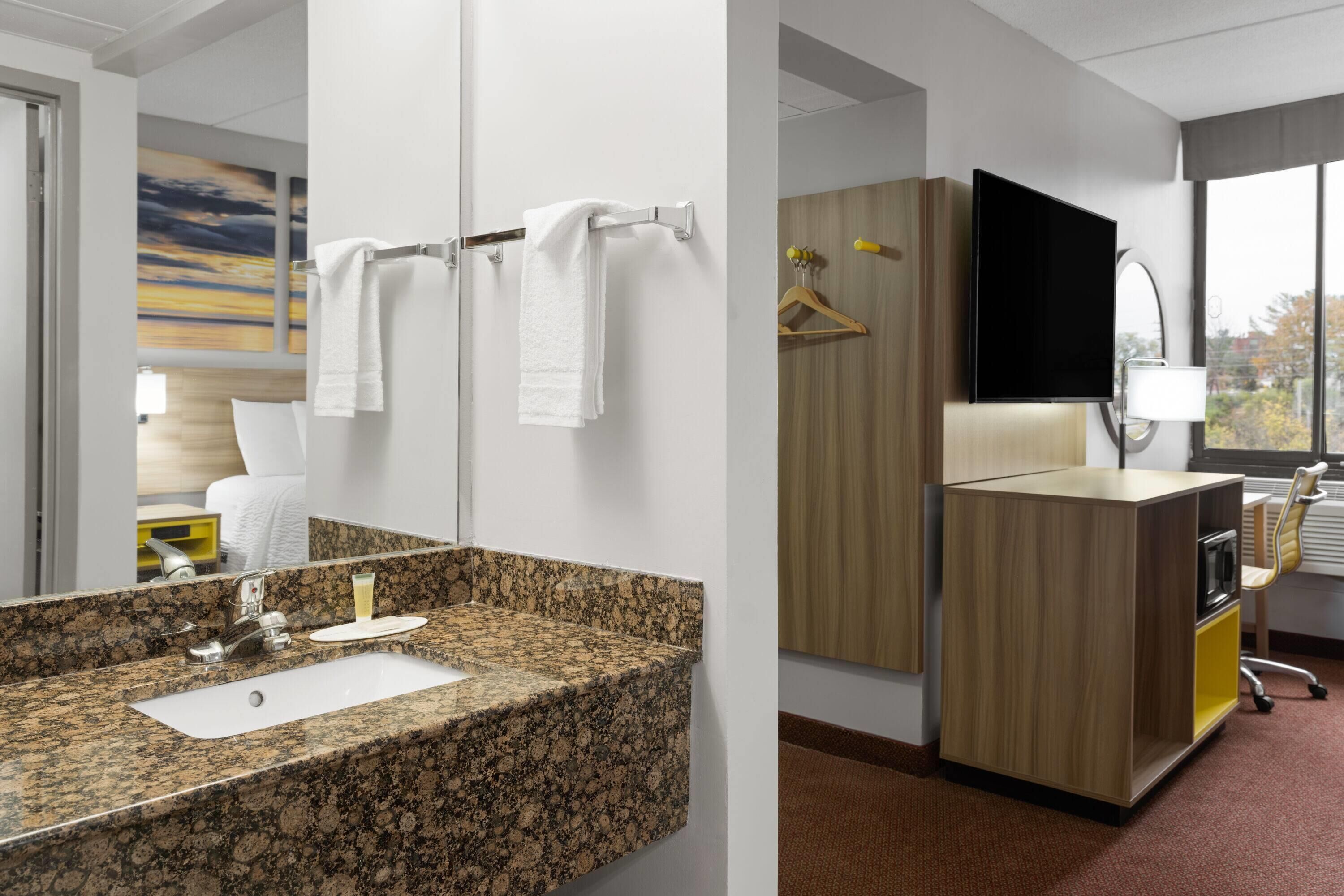 A modern bathroom with a granite countertop sink, a mirror, a towel rack, and a wooden cabinet with a flat-screen TV.