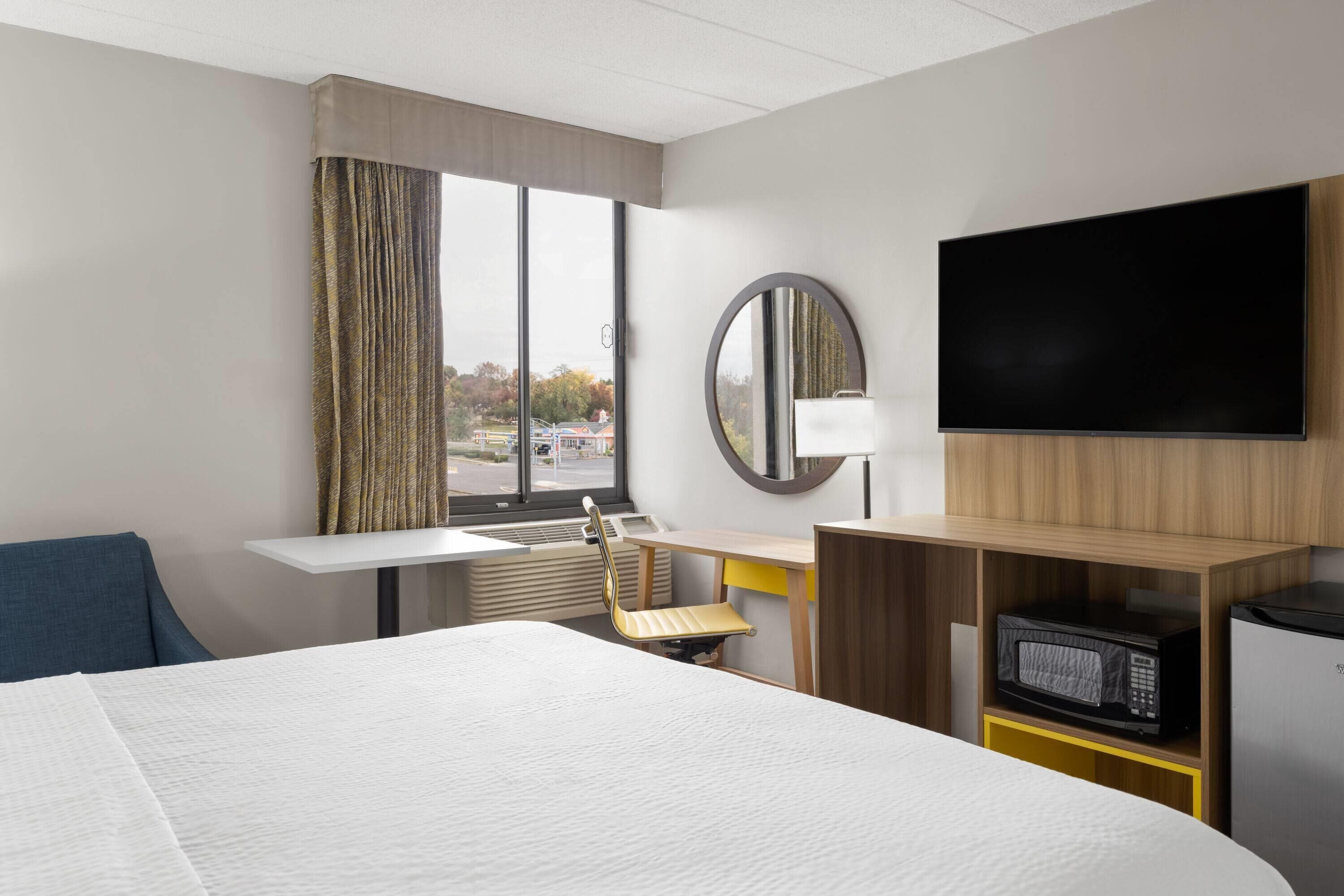 A hotel room with a bed, a desk, a chair, a TV, a microwave, and a view of the outside.