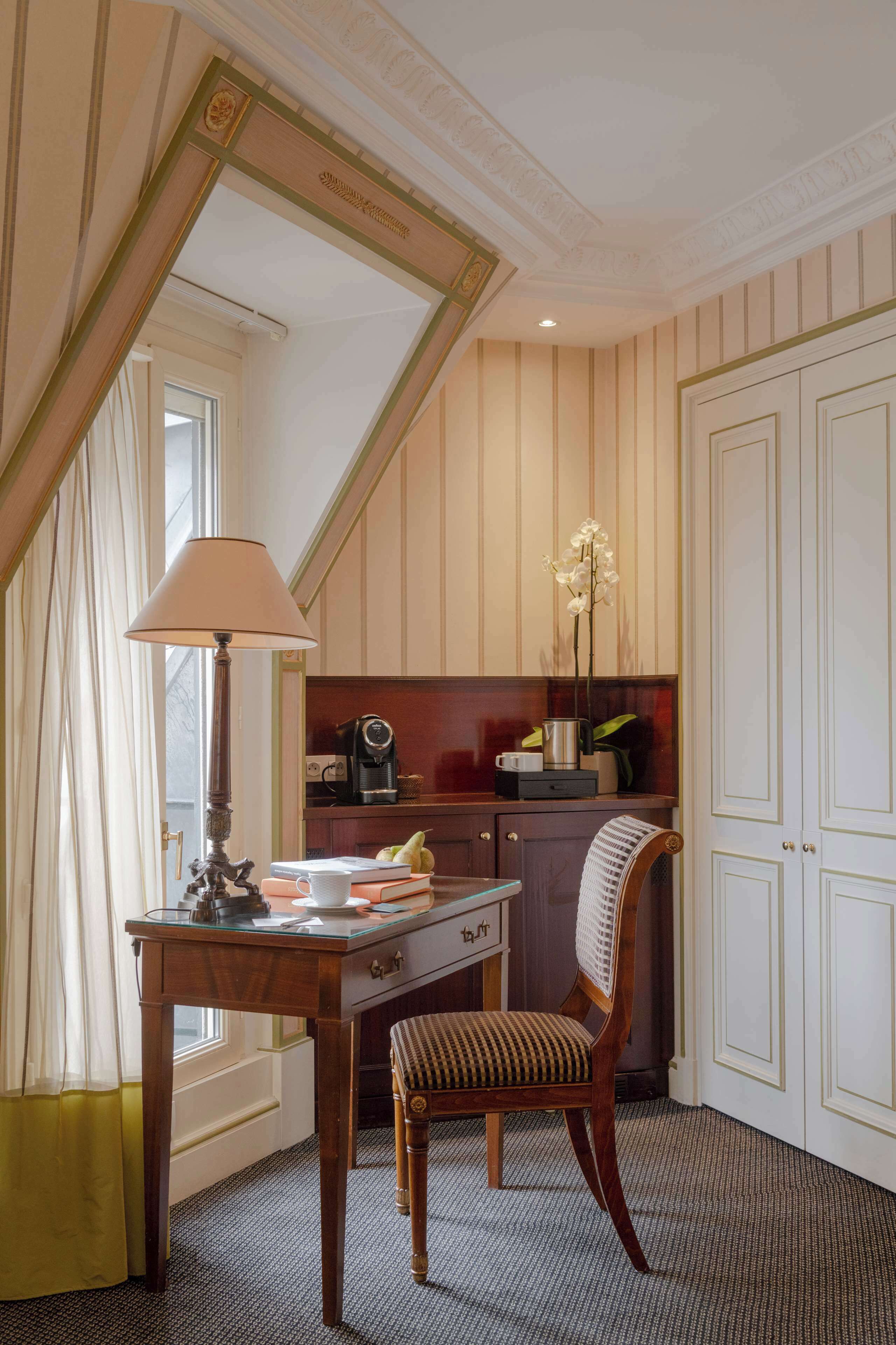 A room with a desk, chair, lamp, and a small kitchenette area.