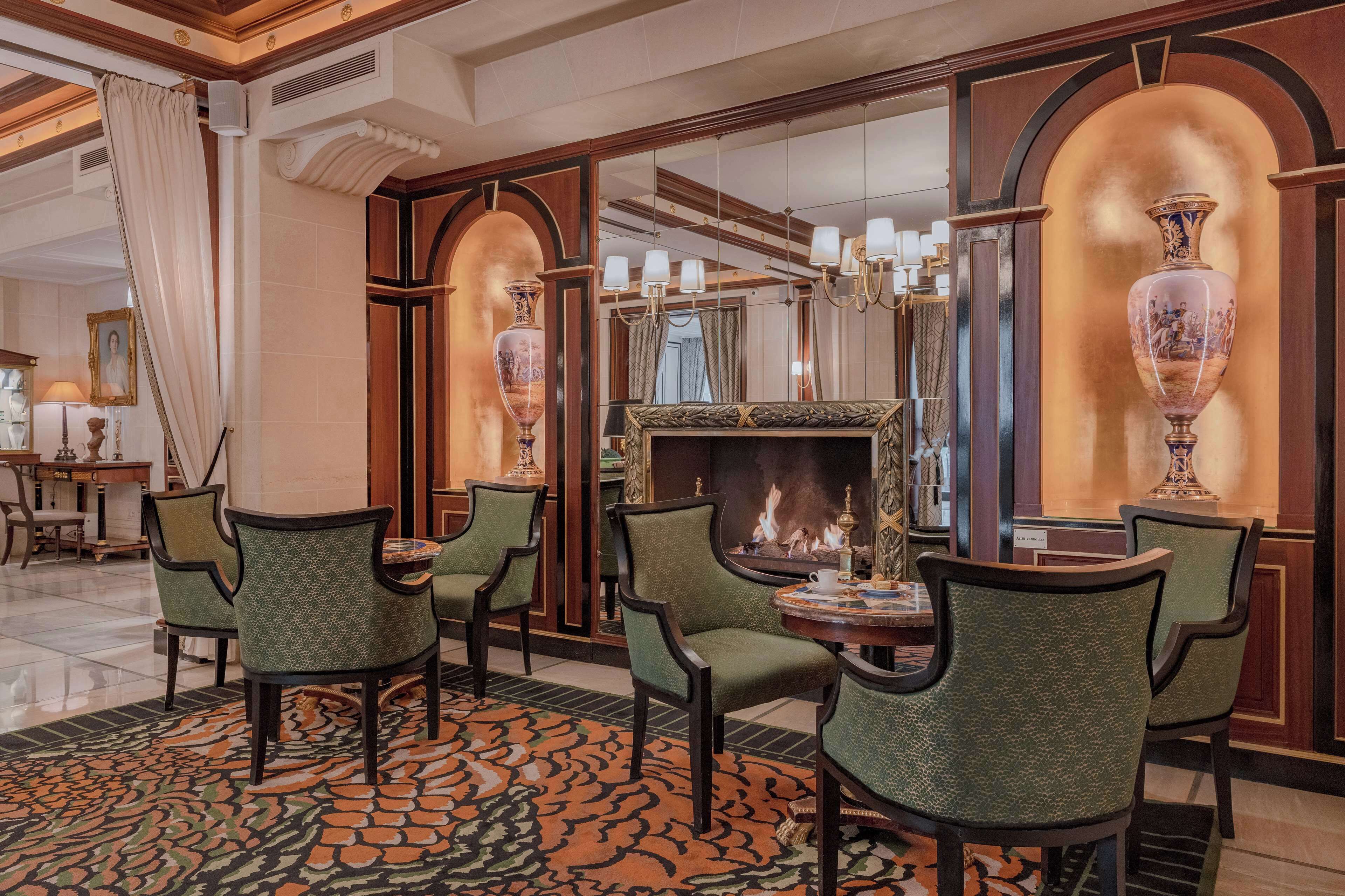 A hotel lobby with a fireplace, green upholstered chairs, a patterned carpet, and a large mirror.