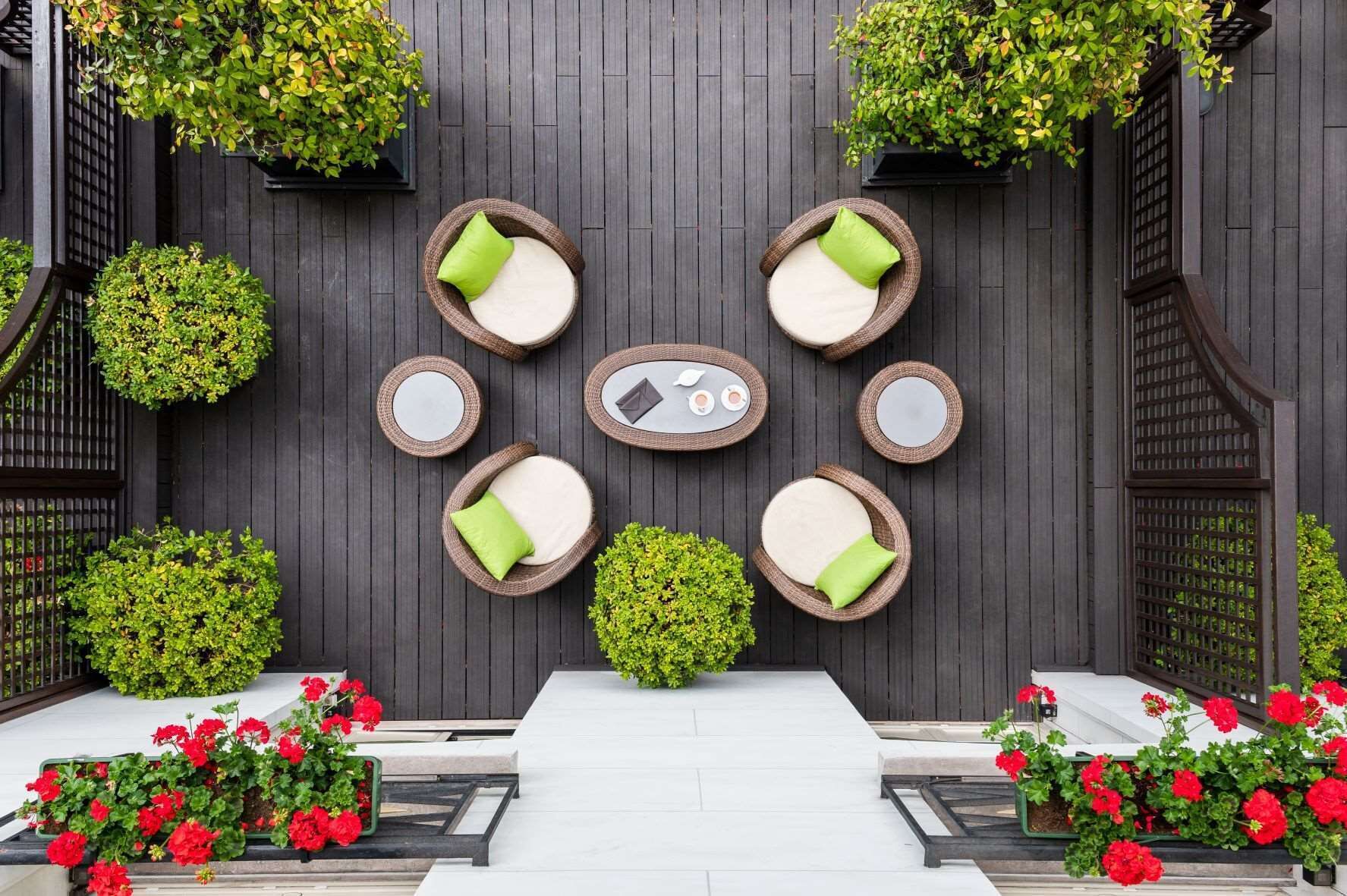 A modern outdoor seating area with round wicker tables and white cushions, surrounded by potted plants and red flowers.