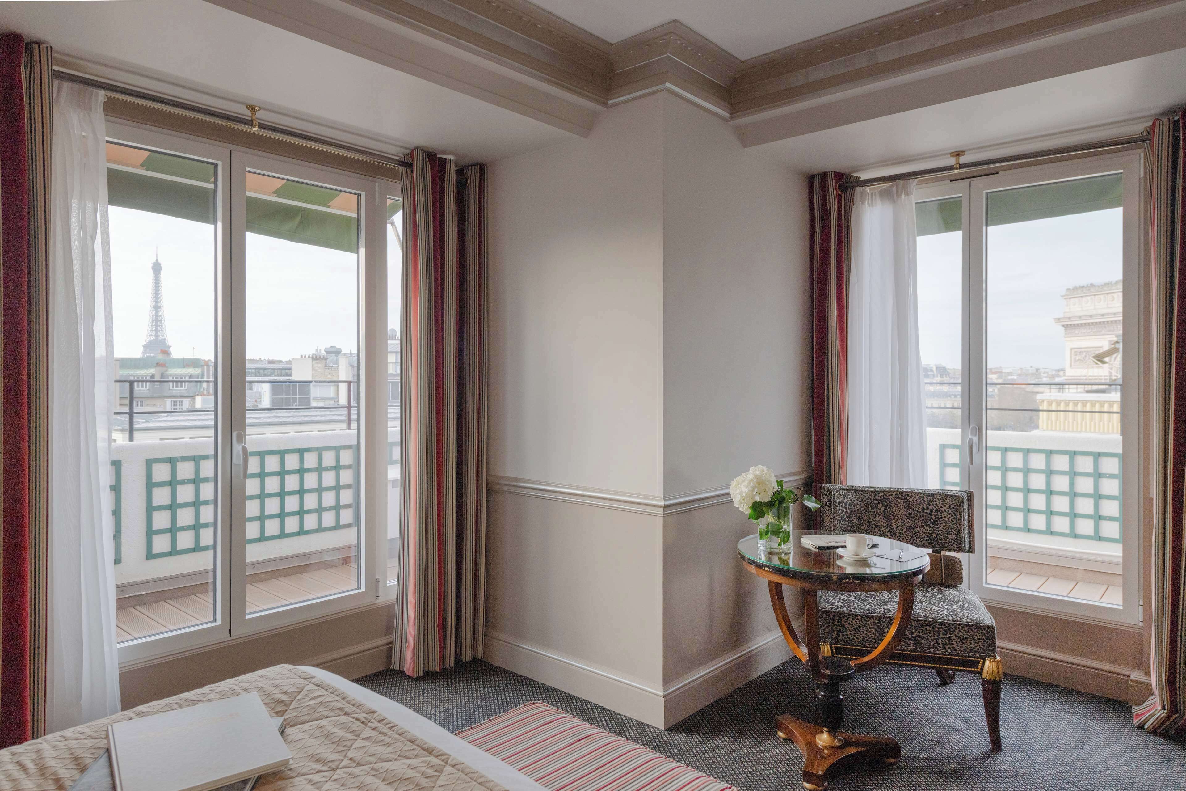 A hotel room with a bed, a small table with a chair, and large windows offering a view of the Eiffel Tower.