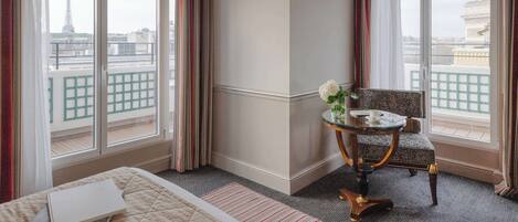 A hotel room with a bed, a small table with a chair, and large windows offering a view of the Eiffel Tower.