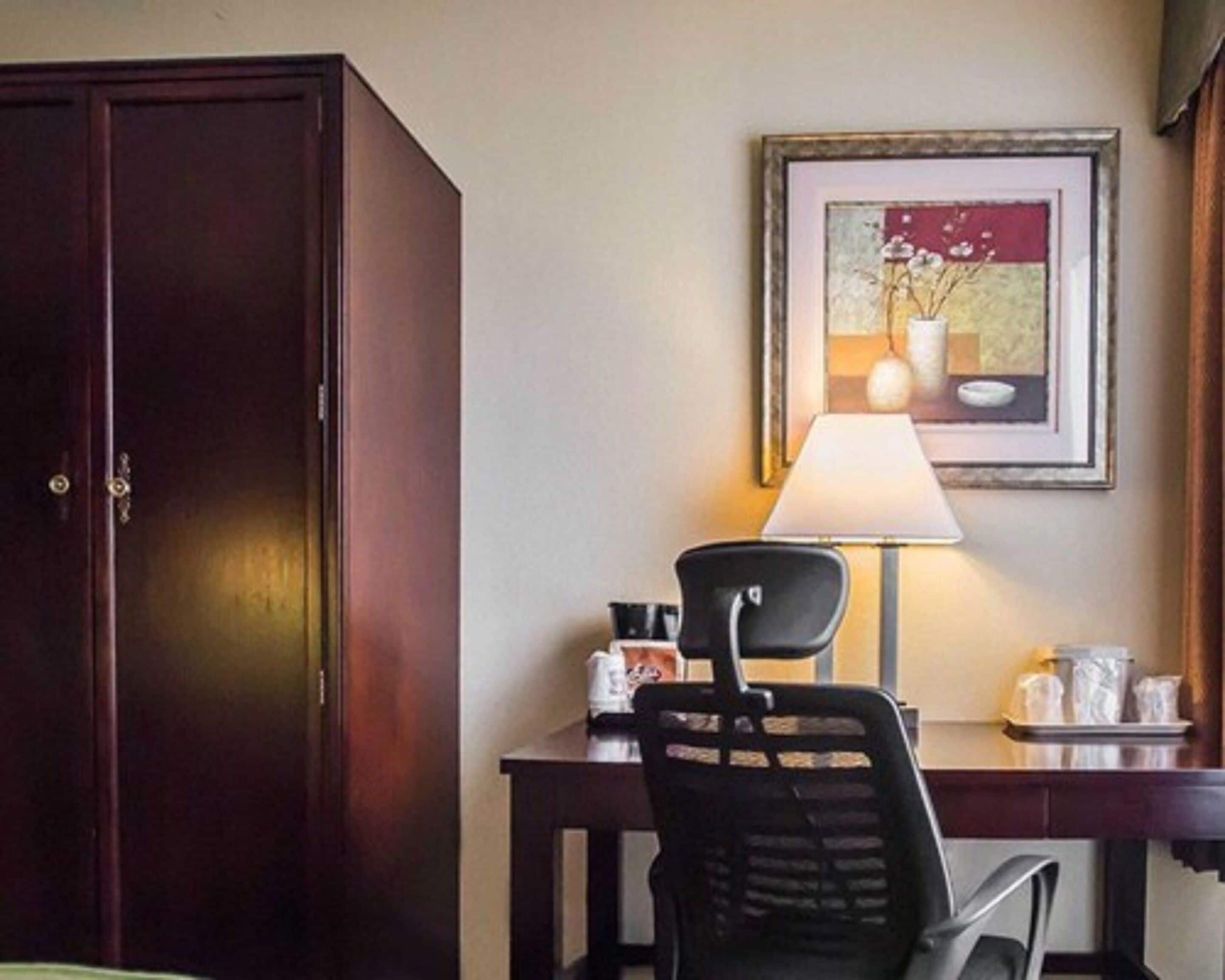 A hotel room with a wooden wardrobe, a desk with a chair, a framed picture on the wall, and a table lamp.