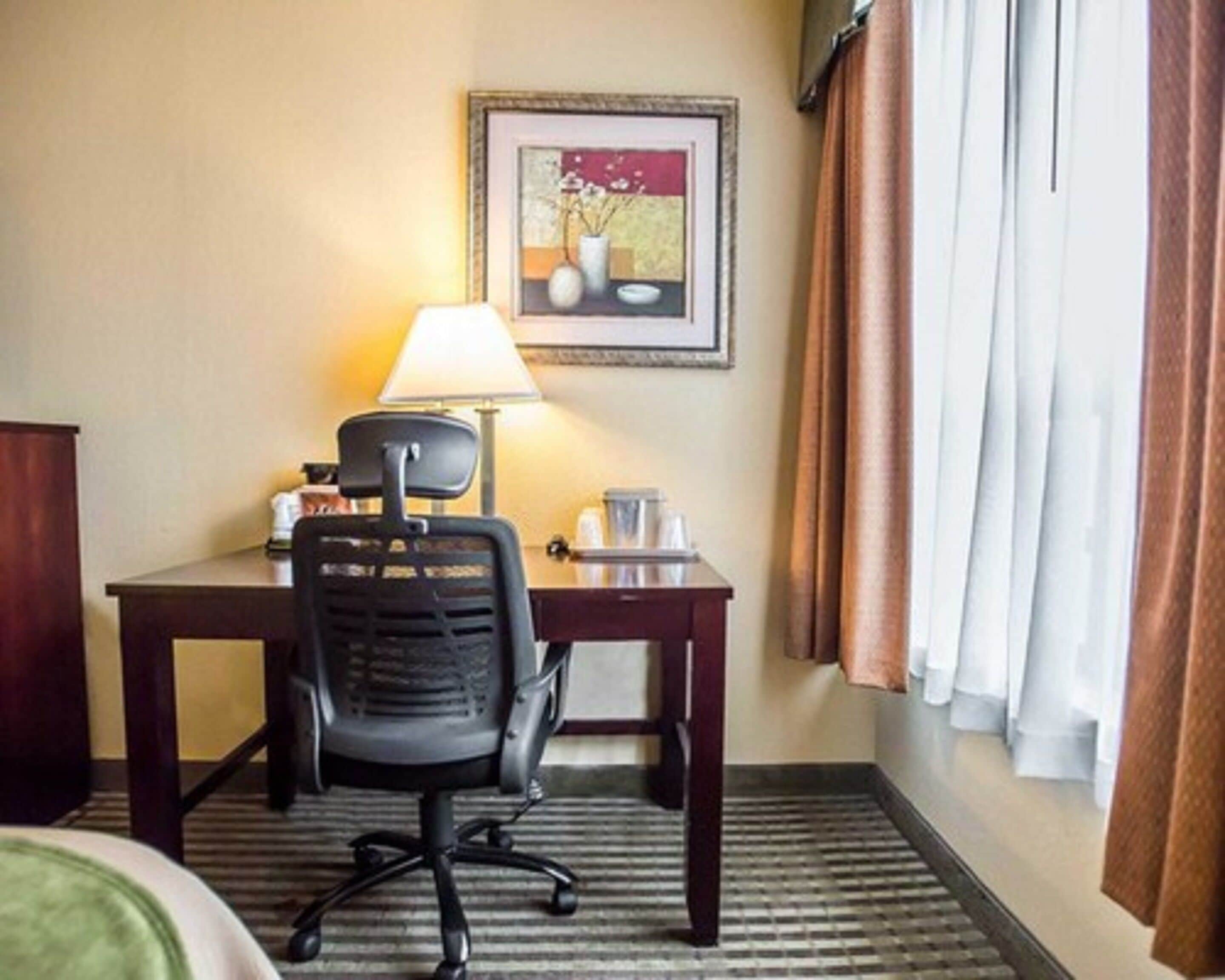 A hotel room with a desk, chair, lamp, framed artwork, and a window with curtains.