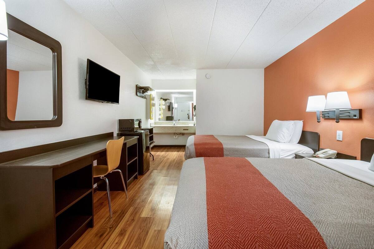 A hotel room with two beds, a desk, a chair, a TV, and a bathroom visible in the background.