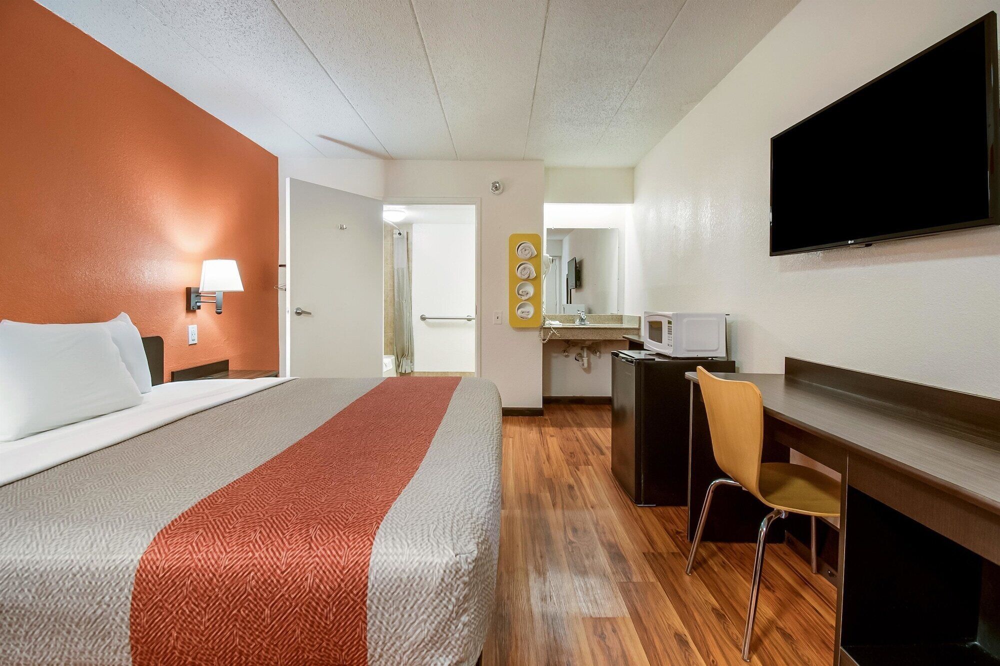 A hotel room with a bed, a desk, a chair, a TV, and a bathroom visible through an open door.