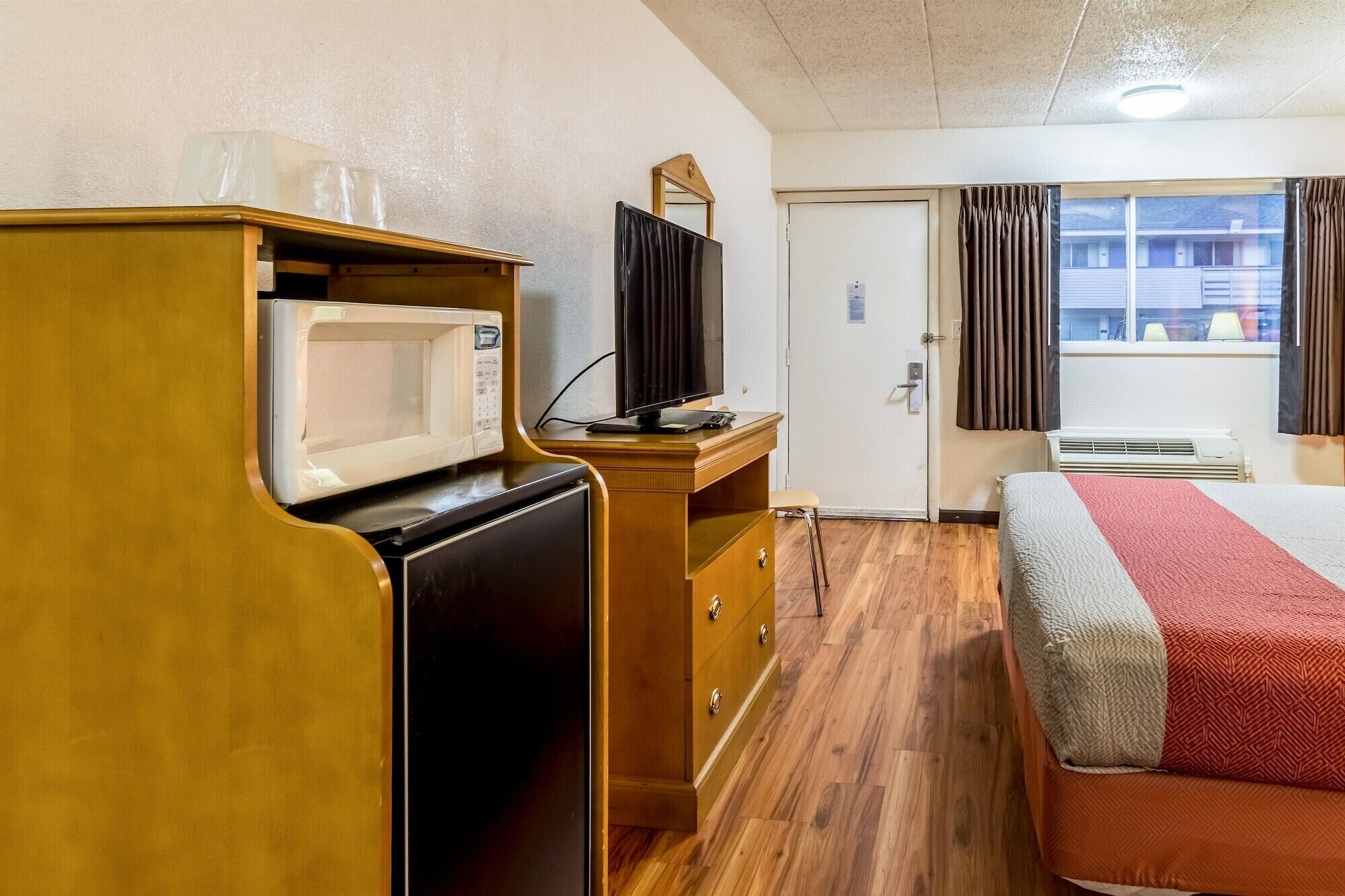 A hotel room with a wooden desk, a microwave, a television, a bed with a red and grey blanket, and a window with curtains.