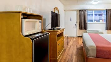 A hotel room with a wooden desk, a microwave, a television, a bed with a red and grey blanket, and a window with curtains.