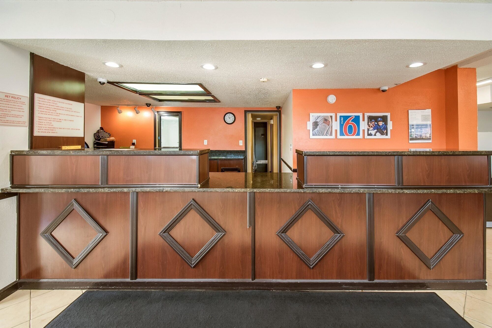 A hotel reception desk with a wooden finish and diamond-shaped panels. There is a clock on the wall and a sign with pet policy information.