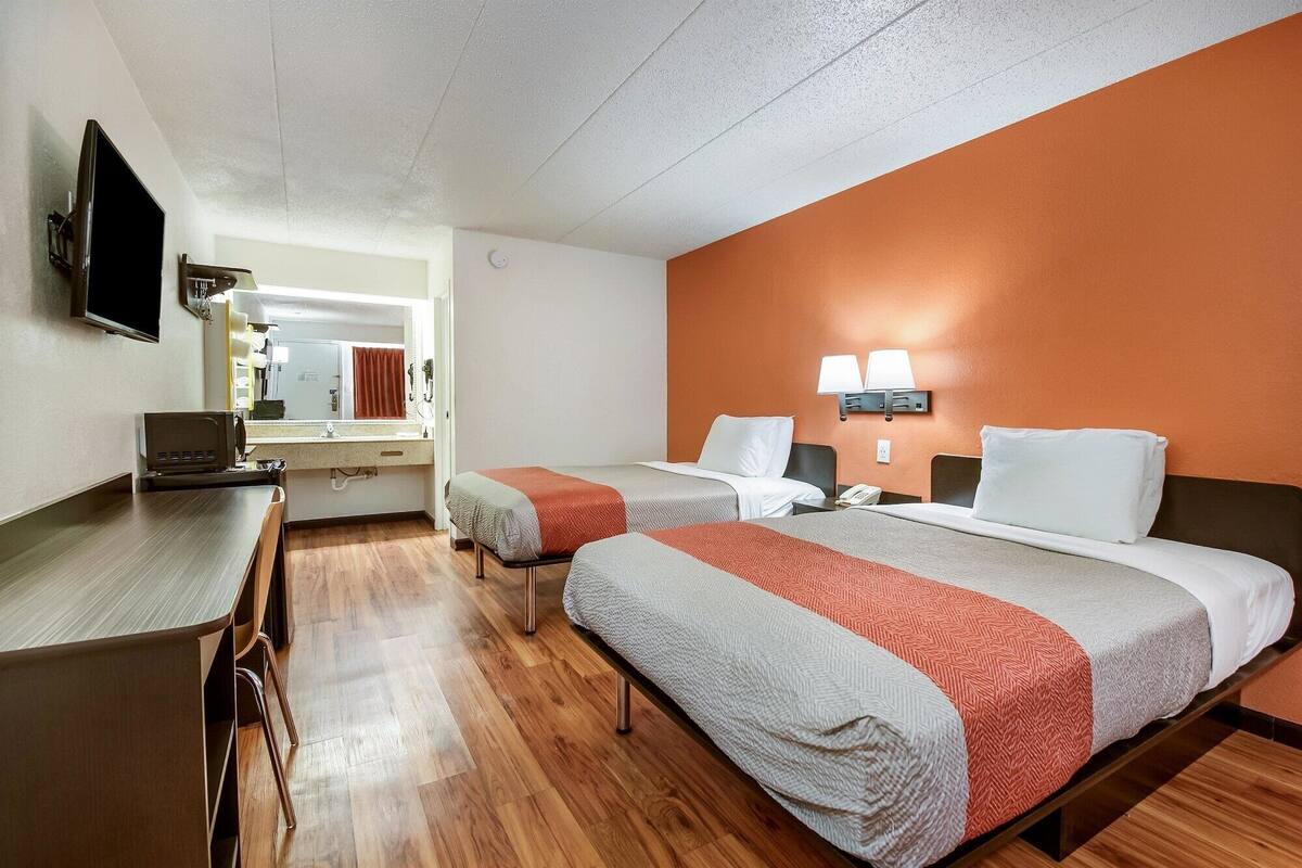 A hotel room with two beds, a desk, a TV, a mirror, and a bathroom visible through an open door.