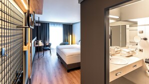 A modern hotel room with a wooden floor, a bed, a desk, and a large mirror in the bathroom.