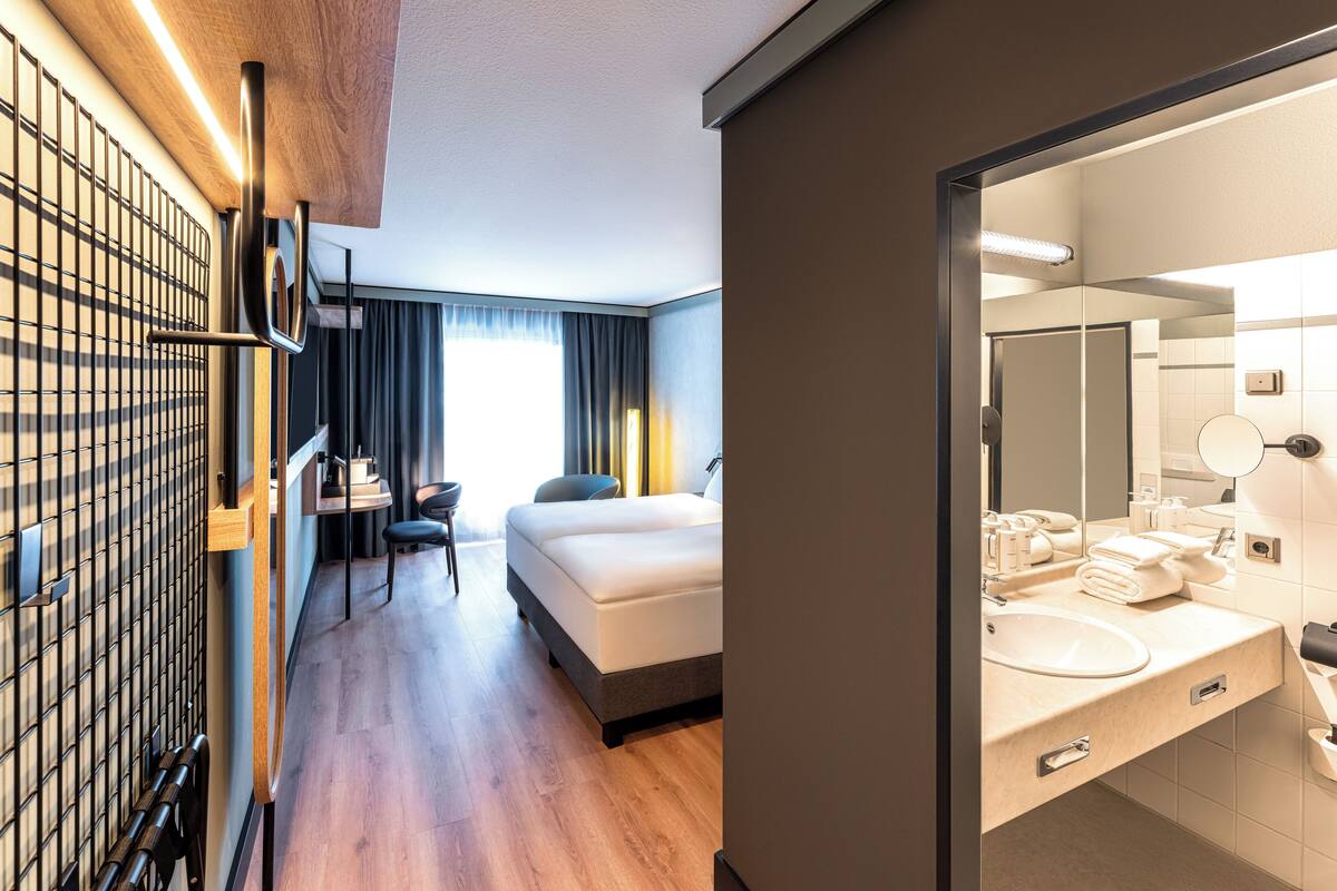 Een moderne hotelkamer met een houten vloer, een bed, een bureau en een grote spiegel in de badkamer.