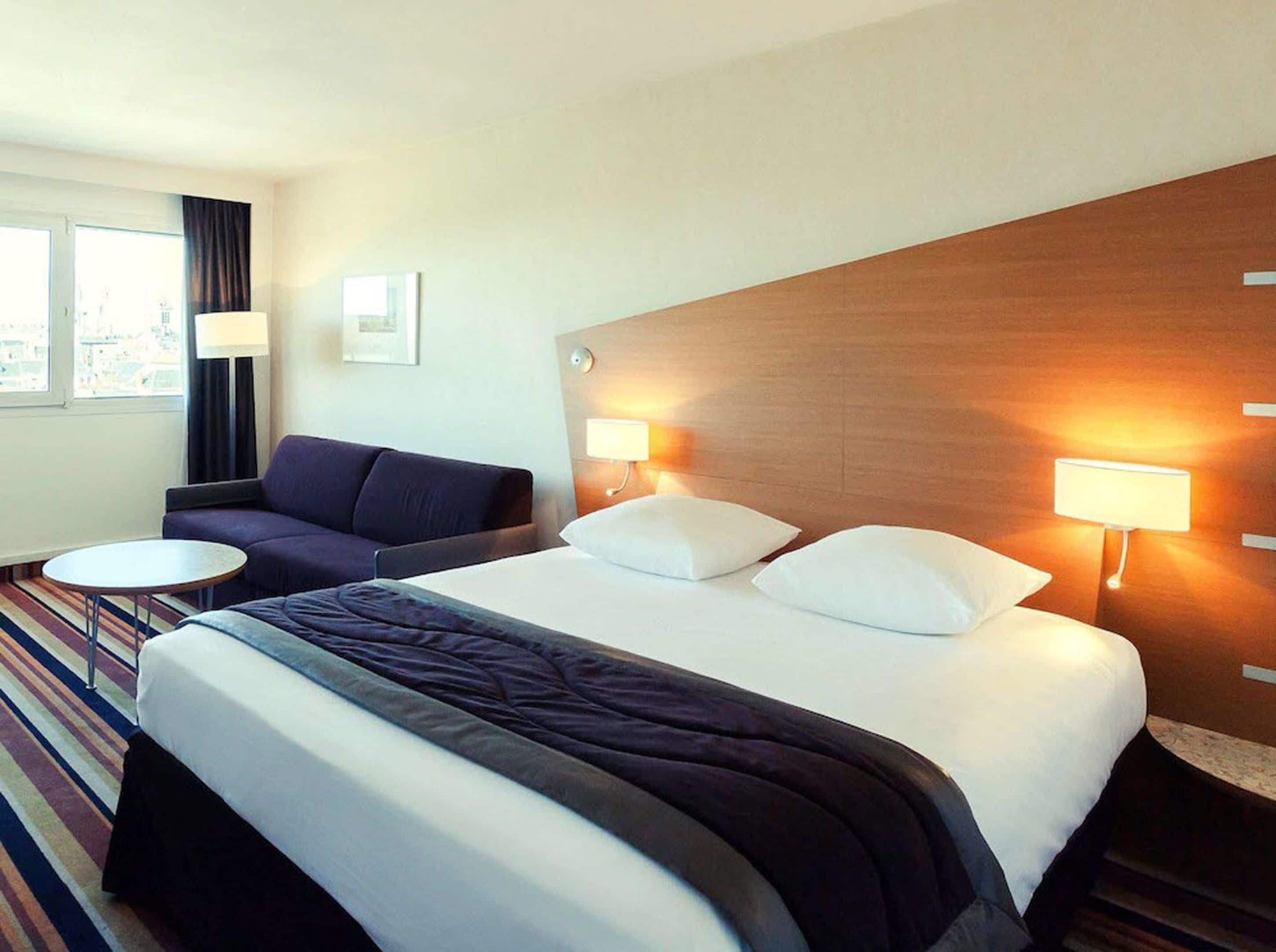 A hotel room with a large bed, two bedside lamps, a sofa, a small round table, and a window with curtains.