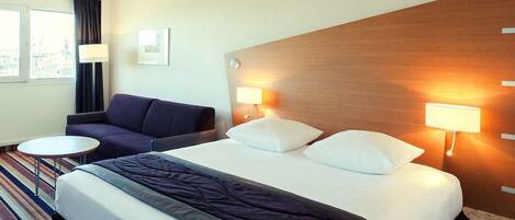 A hotel room with a large bed, two bedside lamps, a sofa, a small round table, and a window with curtains.