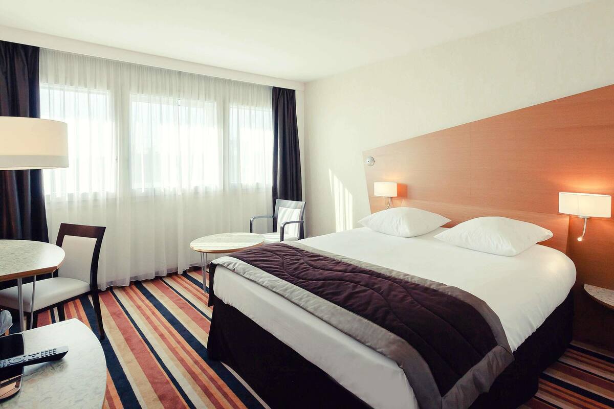 A hotel room with a bed, two bedside lamps, a chair, a small table, and a TV.