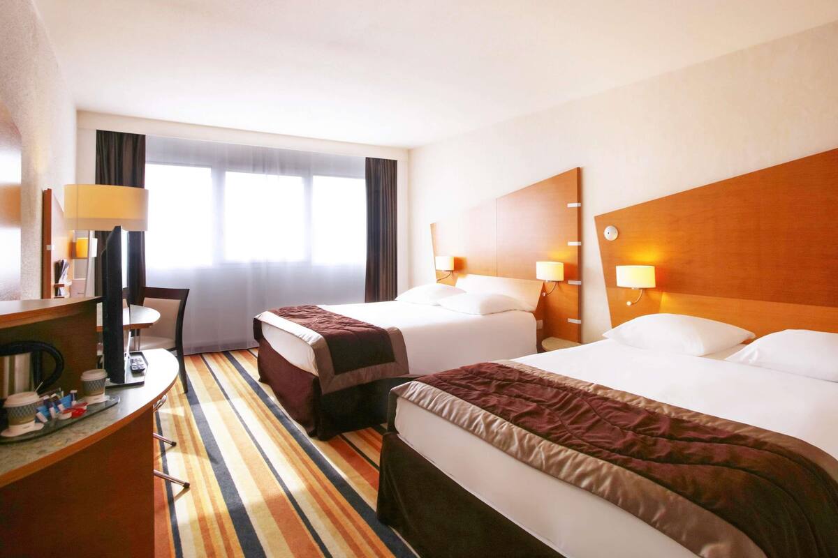 A hotel room with two beds, a desk, a chair, a lamp, and a window with curtains.