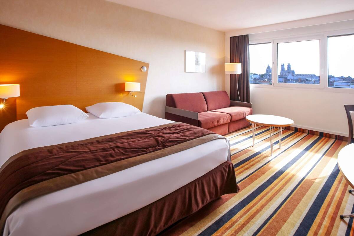 A hotel room with a large bed, a sofa, a small table, and a view of a cityscape through the window.