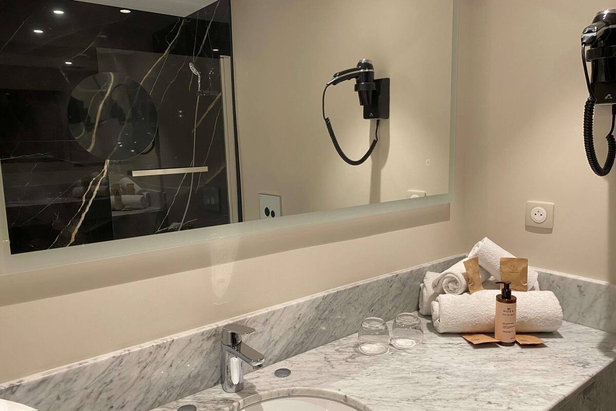 A bathroom with a marble countertop, a sink, a mirror, a hairdryer, and toiletries.