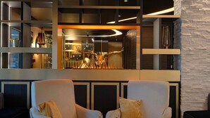 Un área de descanso moderna con dos sillones, una mesita redonda y una zona de bar visible a través de un panel de vidrio.