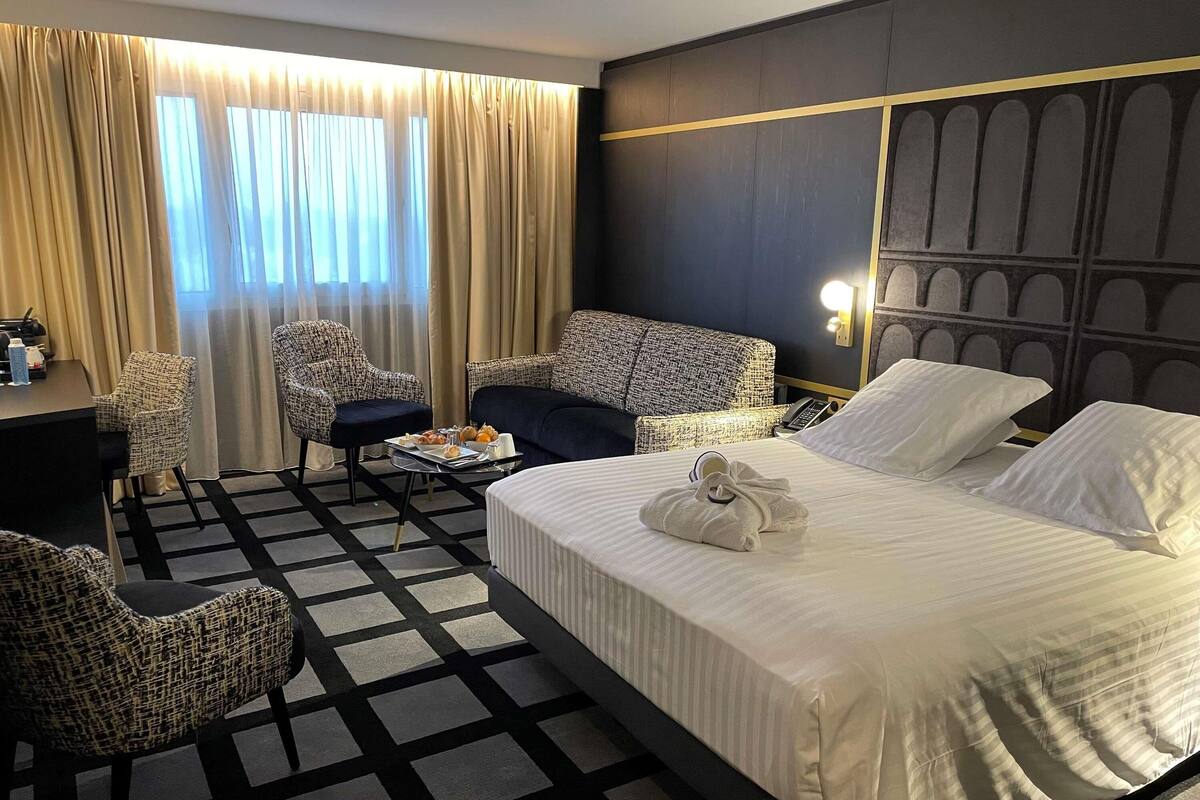 A hotel room with a bed, two armchairs, a desk, and a window with curtains.
