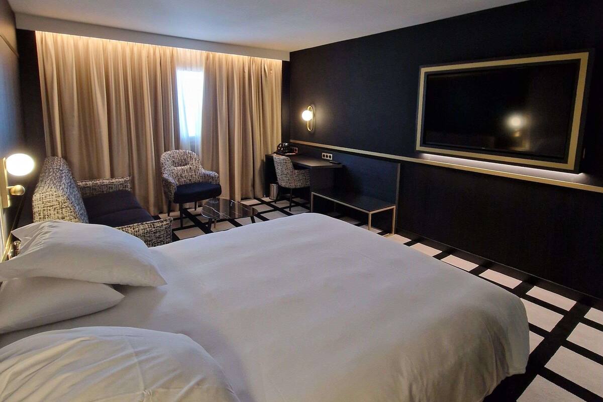 A hotel room with a large bed, a desk, a chair, a TV, and a window with curtains.