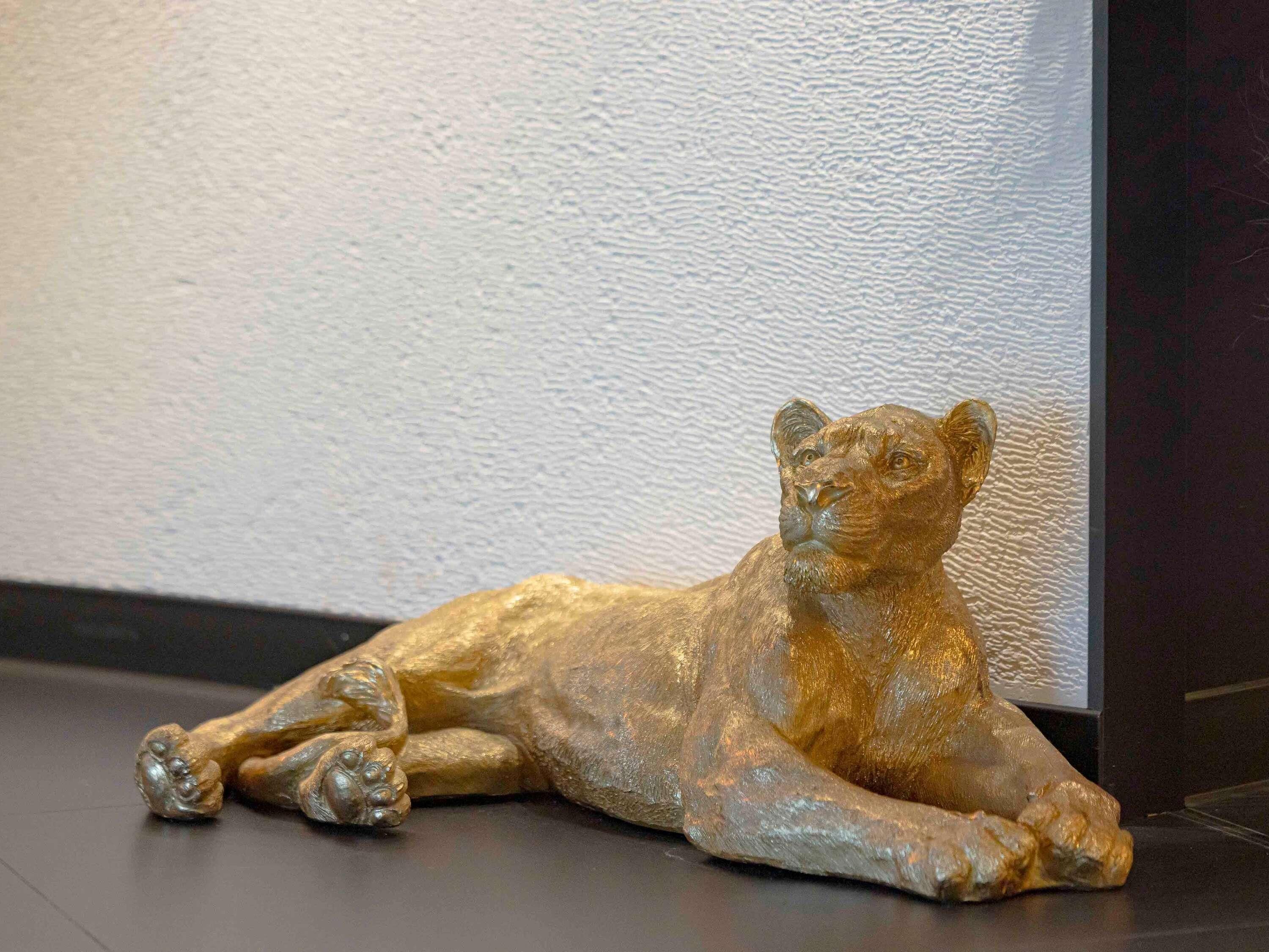 Une statue de lion doré, allongée sur un sol sombre, adossée à un mur à la texture marquée.