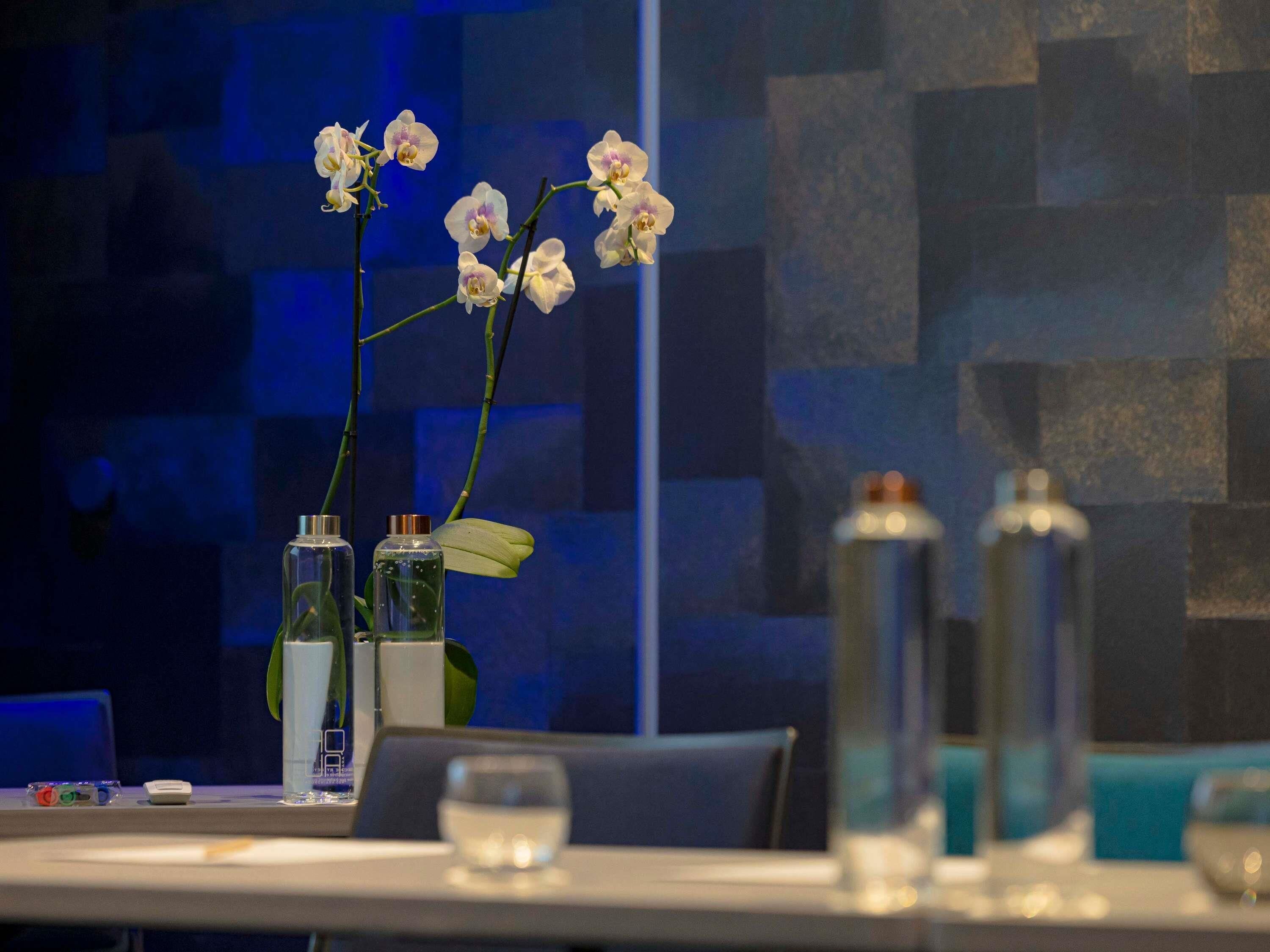A table with vases, a glass of water, and a blue wall with a geometric pattern.