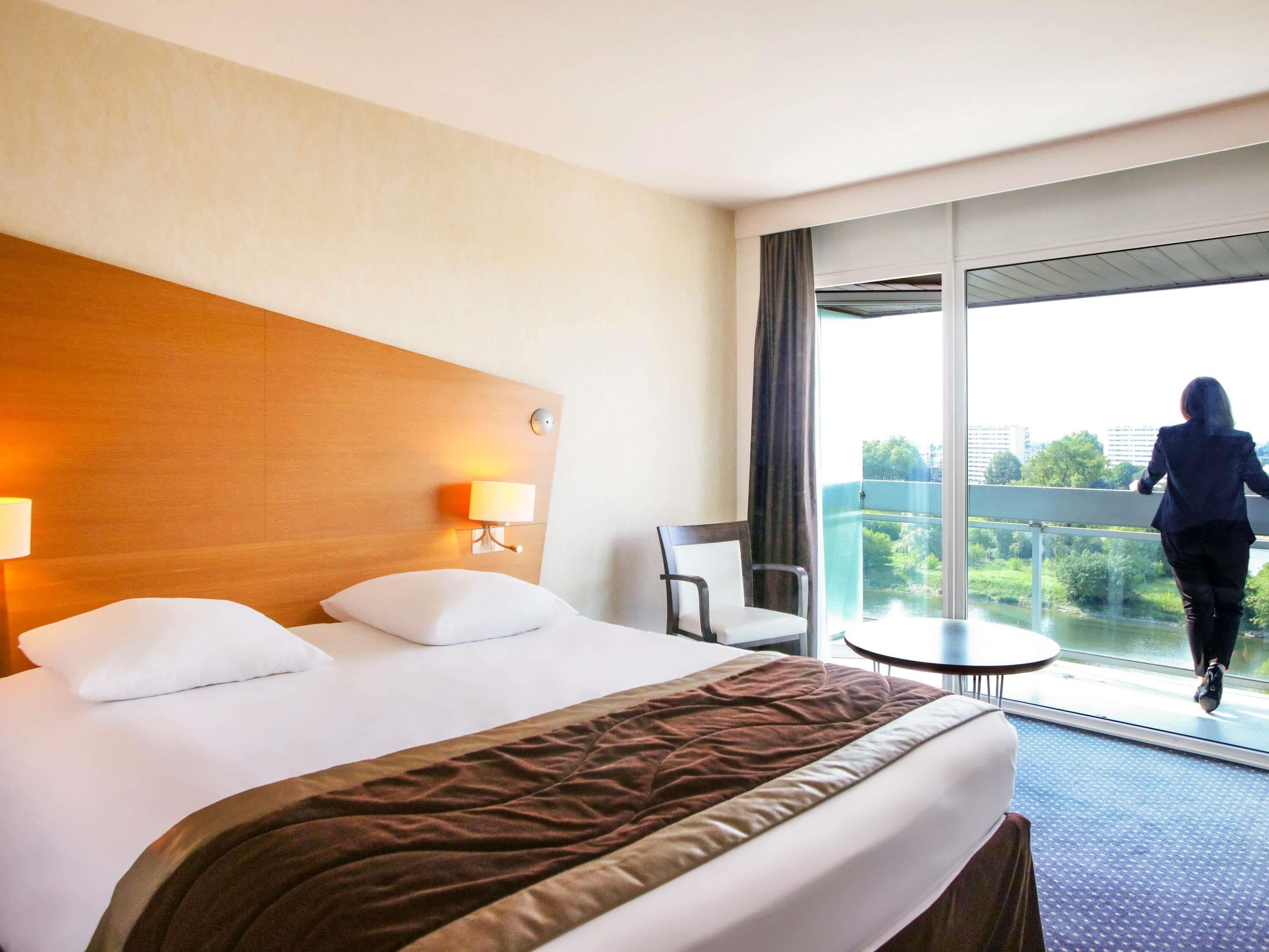 Una habitación de hotel con cama, lámpara de noche, una silla, una mesita y vistas a un balcón.