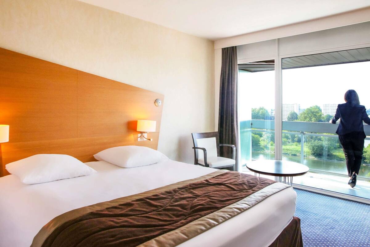 A hotel room with a bed, a nightstand, a lamp, a chair, and a balcony with a view of a river and buildings.