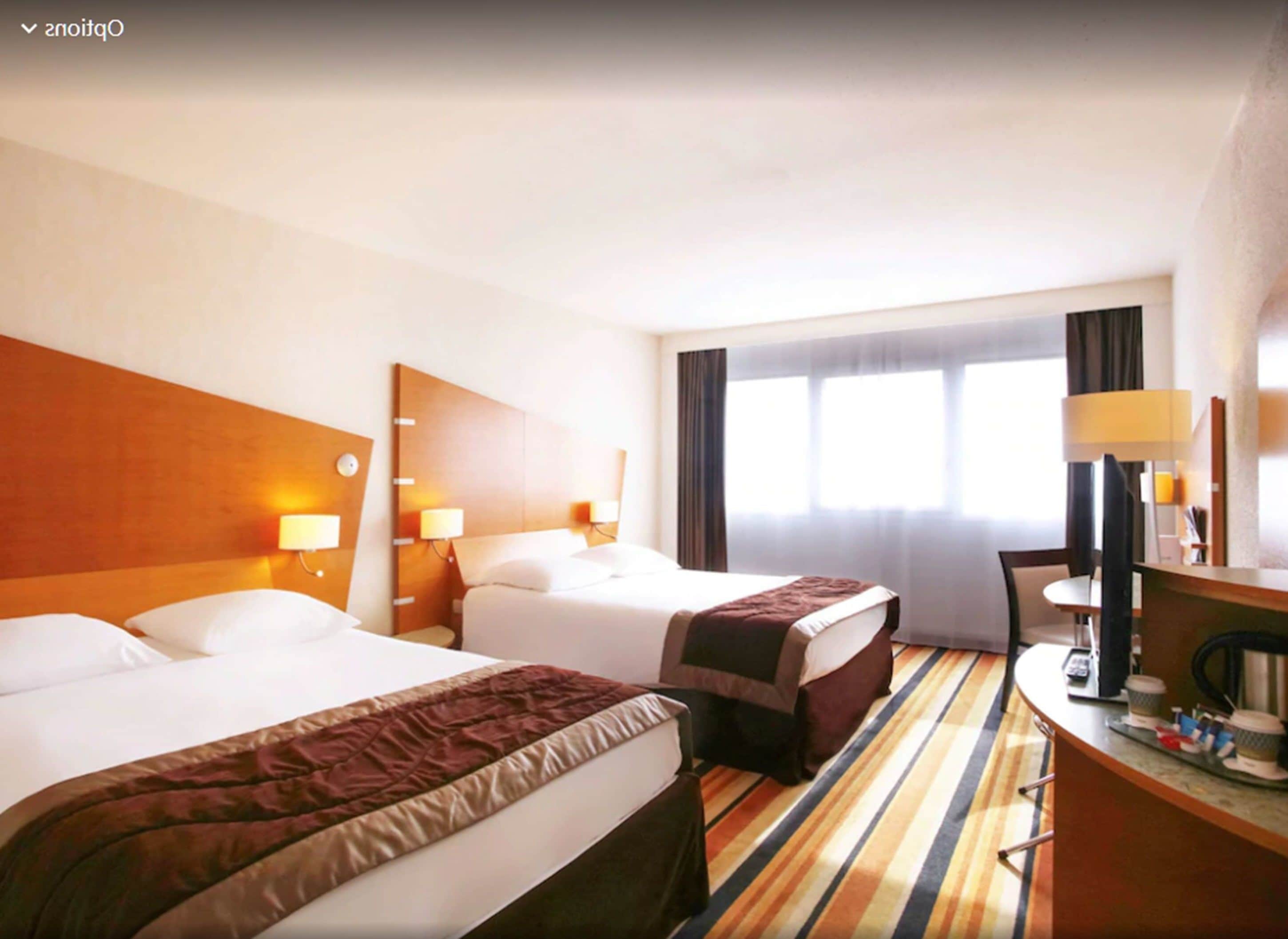 Une chambre d’hôtel avec deux lits, un bureau, une chaise et une fenêtre avec des rideaux.