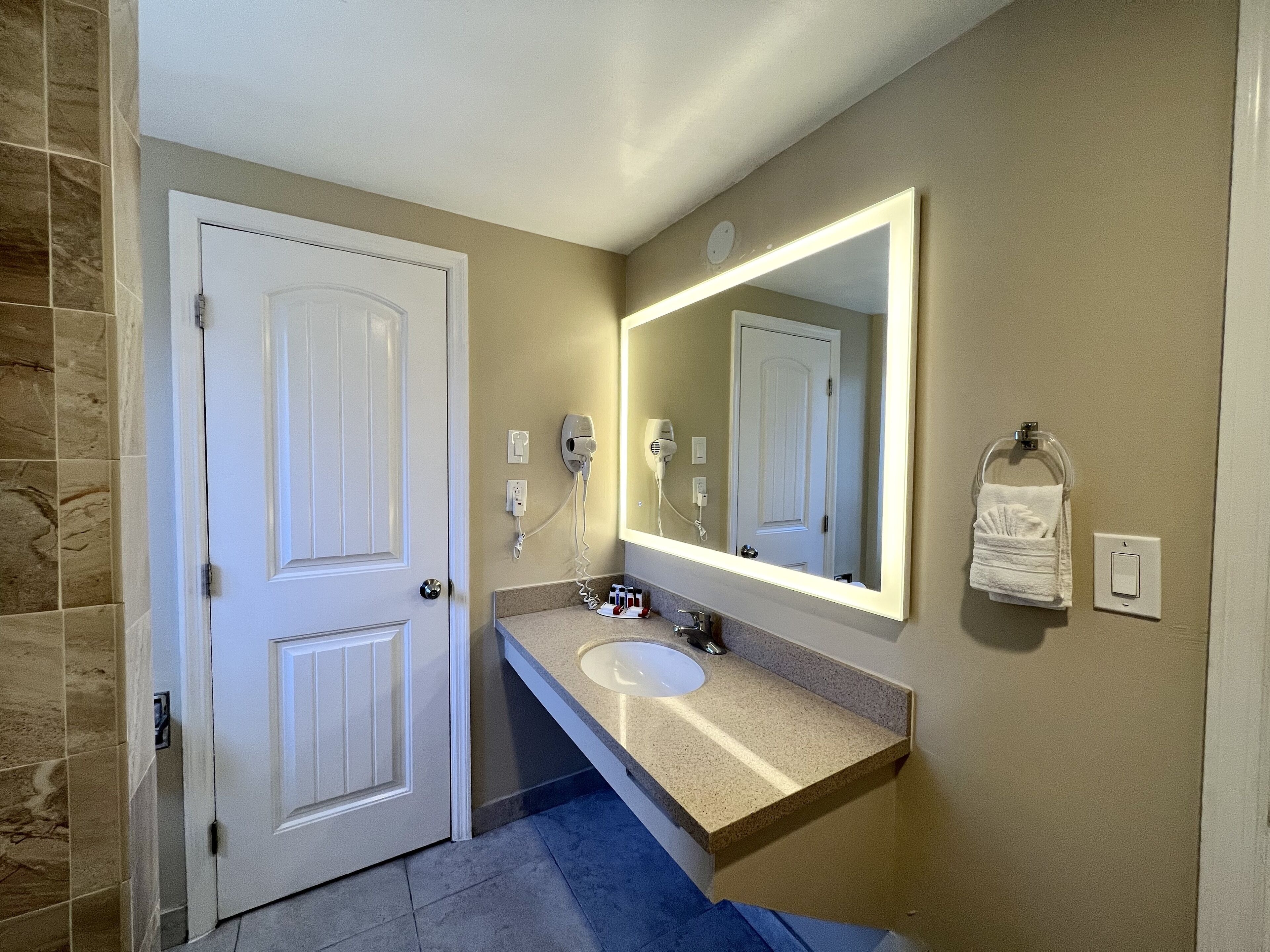 A bathroom with a white door, a large mirror, a sink, and a towel rack.