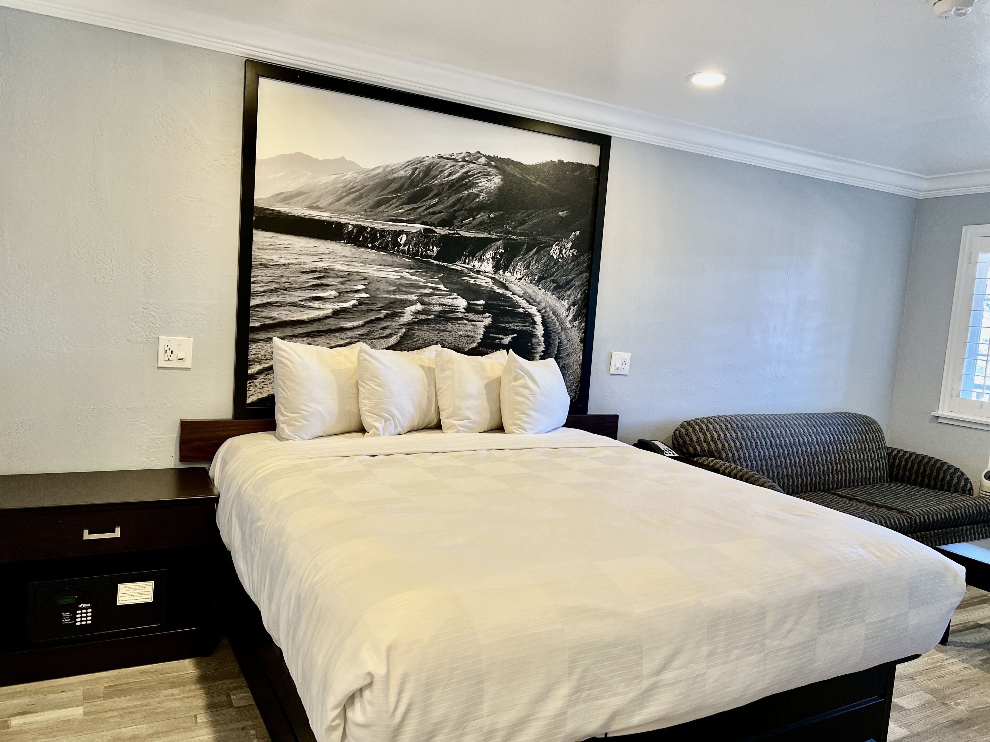 A hotel room with a large bed, a dark wooden nightstand, a black chair, and a large black and white mountain landscape painting on the wall.