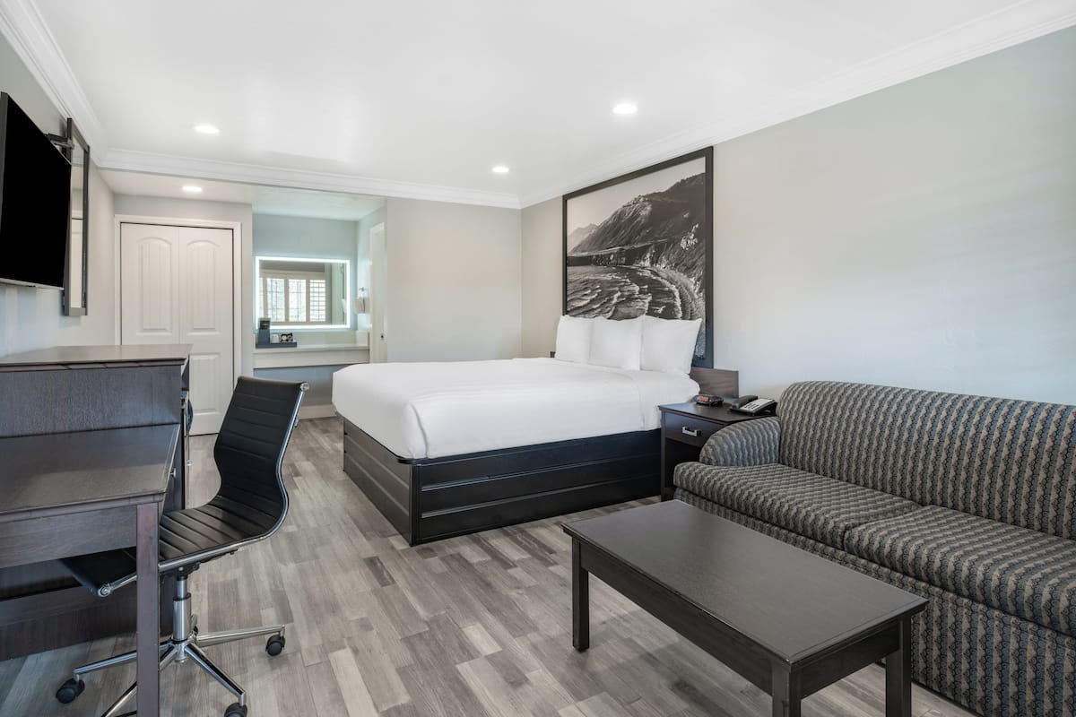 A hotel room with a bed, a sofa, a desk with a chair, a TV, and a painting on the wall.