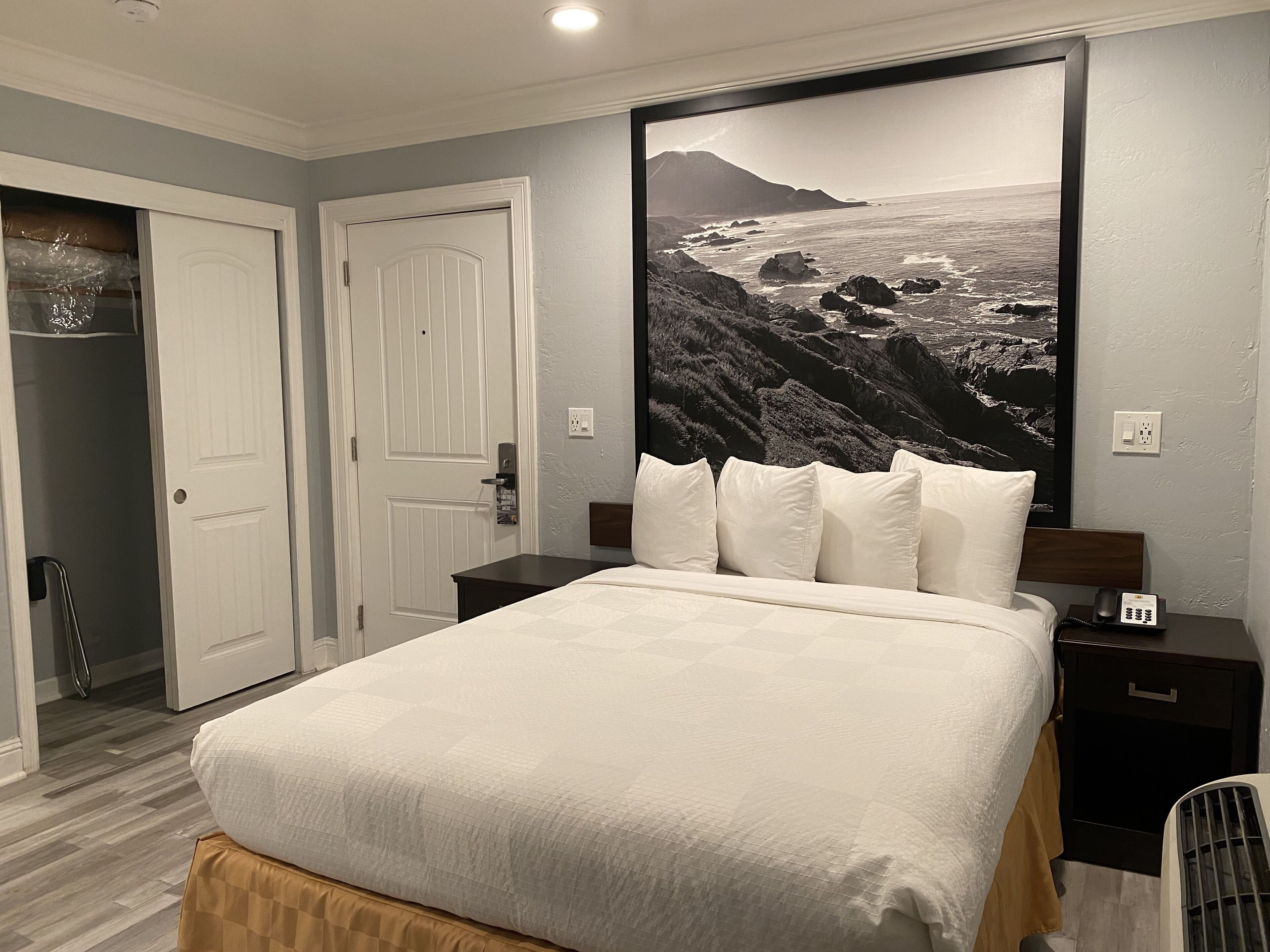Un dormitorio con una cama, una mesita de noche, un teléfono instalado en la pared y un cuadro grande con un paisaje costero.