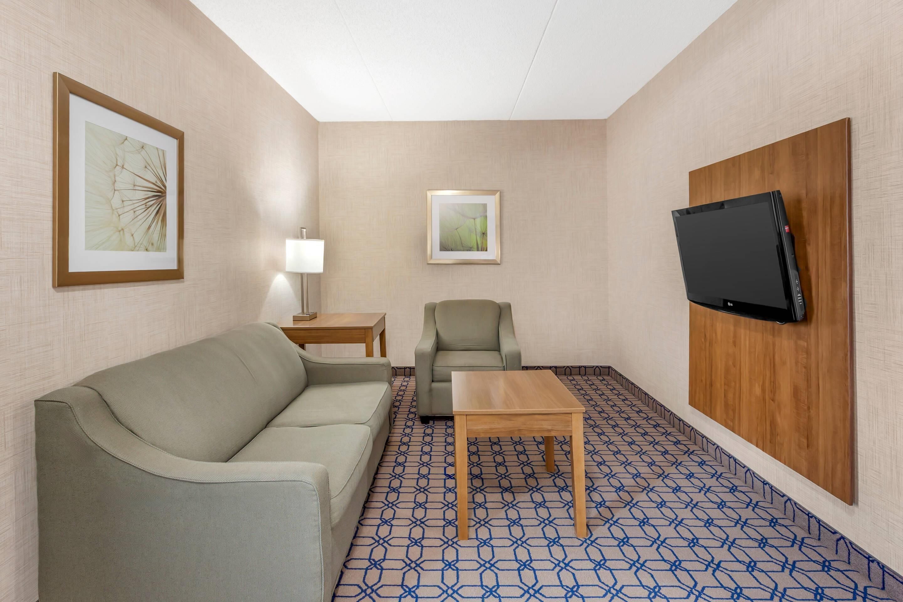 A hotel room with a sofa, a chair, a small table, a TV mounted on the wall, and two framed pictures on the walls.